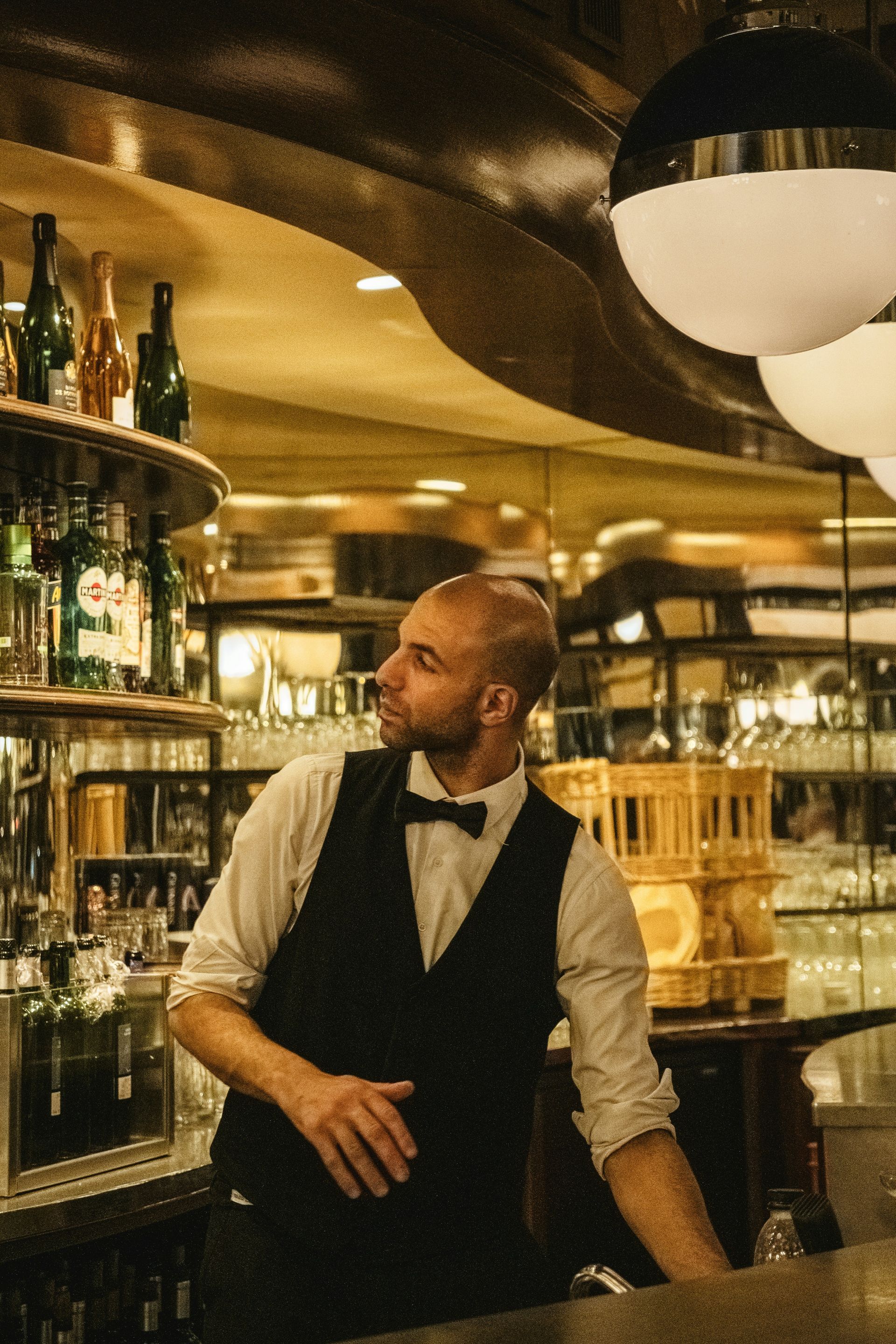 A bald bartender wearing a black vest and bow tie looks towards the side. Behind him, shelves of bottles are visible.