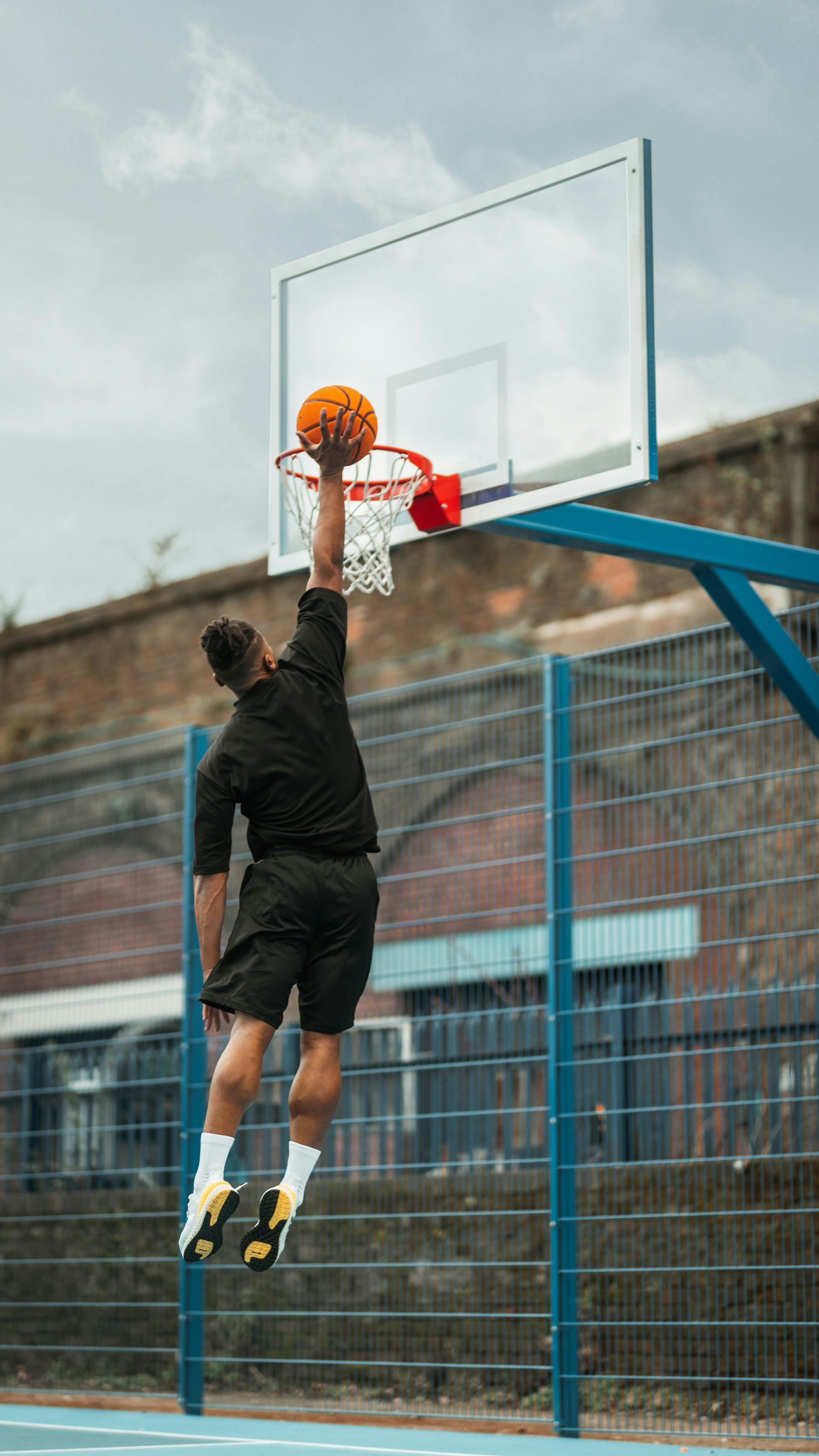 Basketball player jumping for a dunk. Orange ball, black shorts, black shirt, outdoor court.