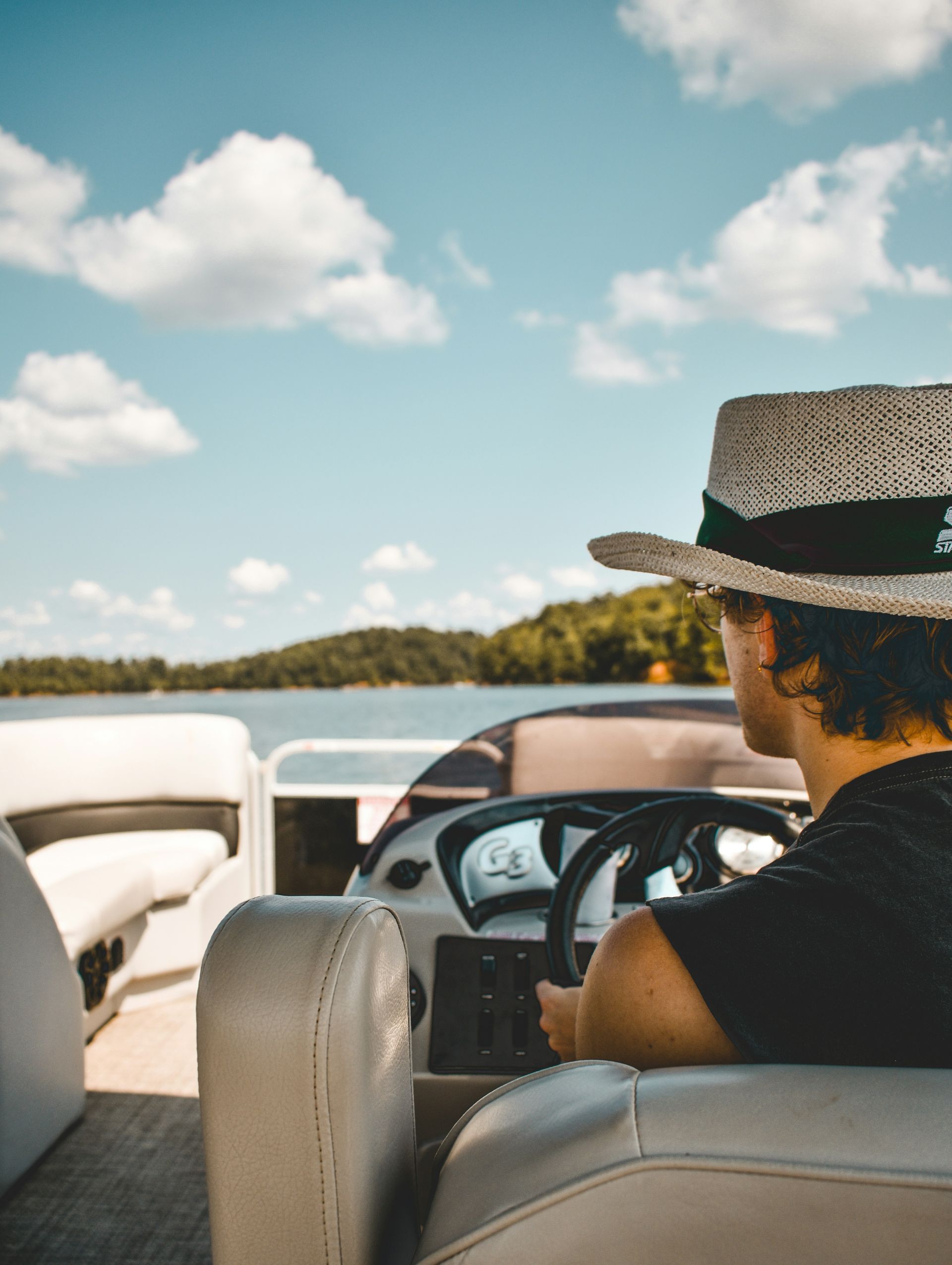 Person steering a pontoon boat on a lake with a blue sky and clouds.