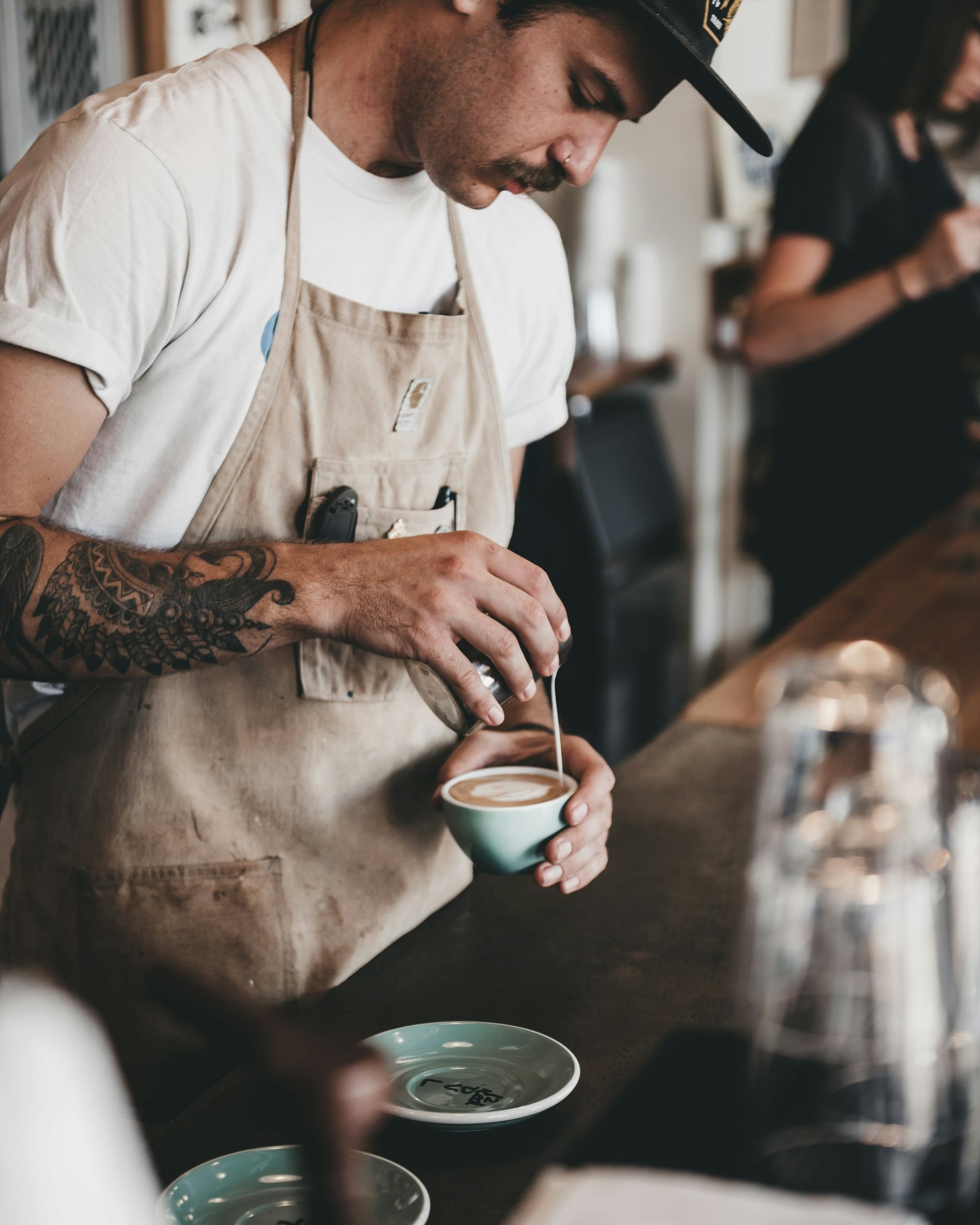 Barista in apron pouring latte art into a small cup, café setting.