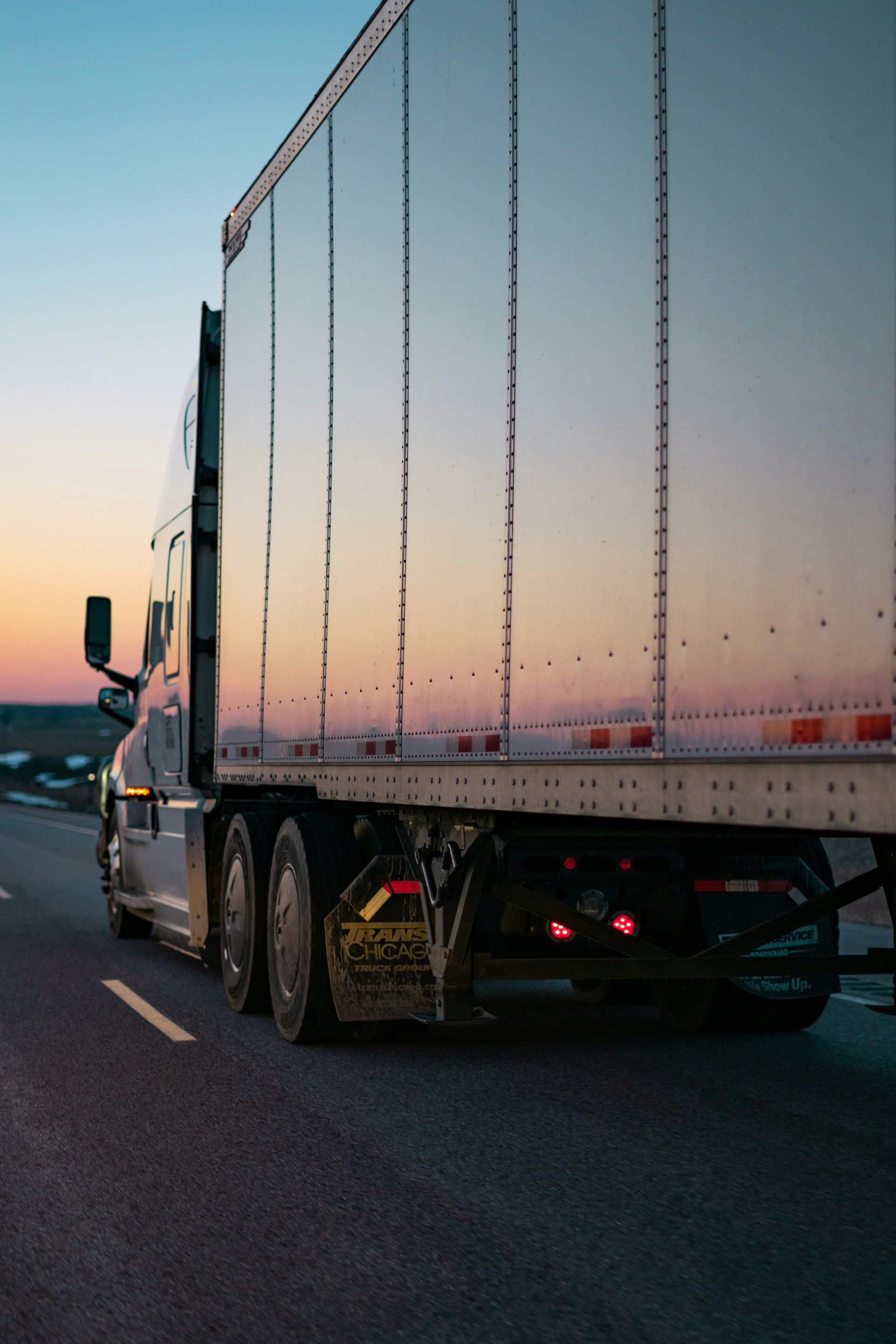 Semi-truck driving on a road at sunset, with a reflective trailer and the sky in shades of orange and blue.