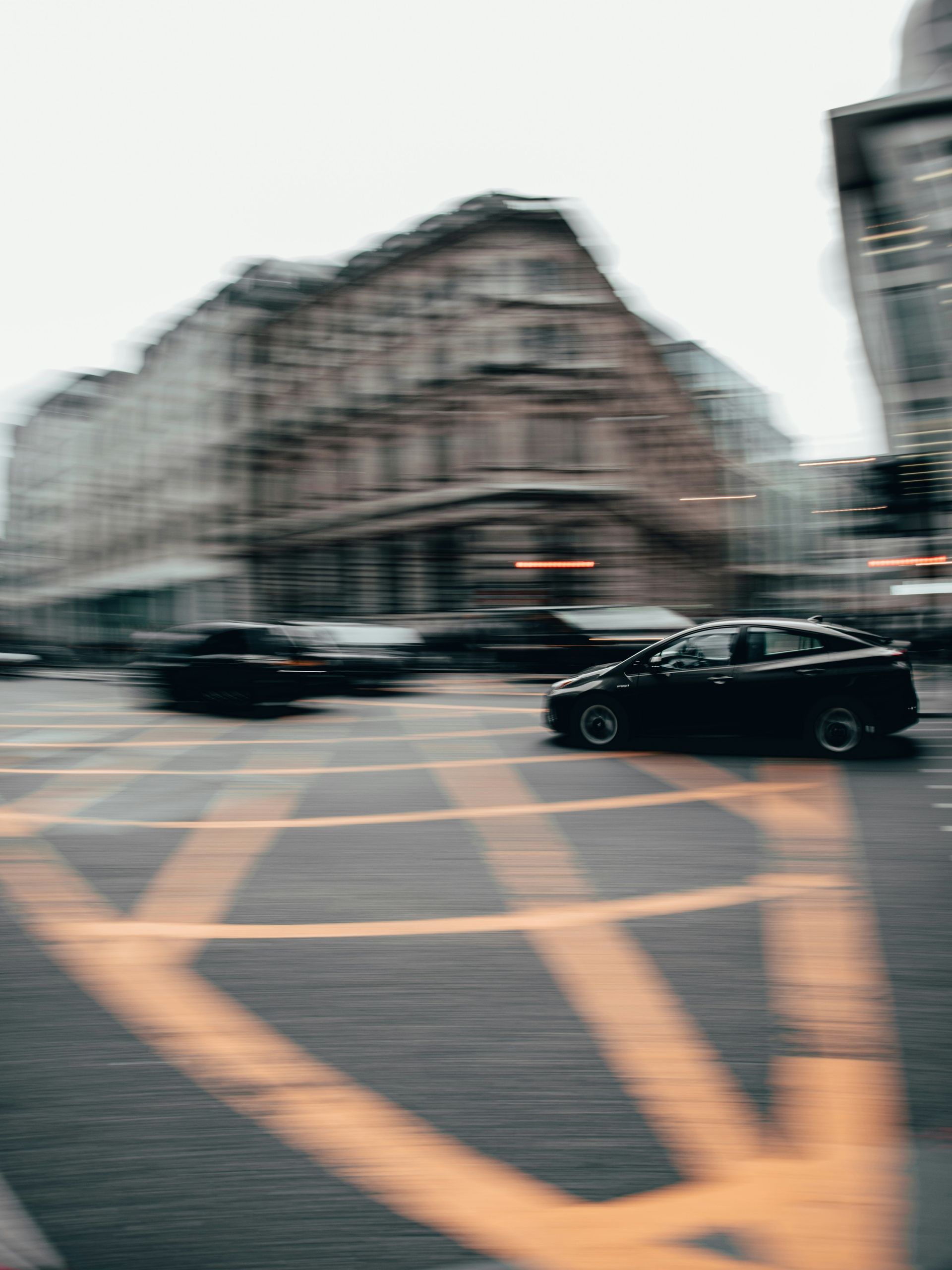Black car in motion crossing a city intersection with blurred yellow markings and buildings.