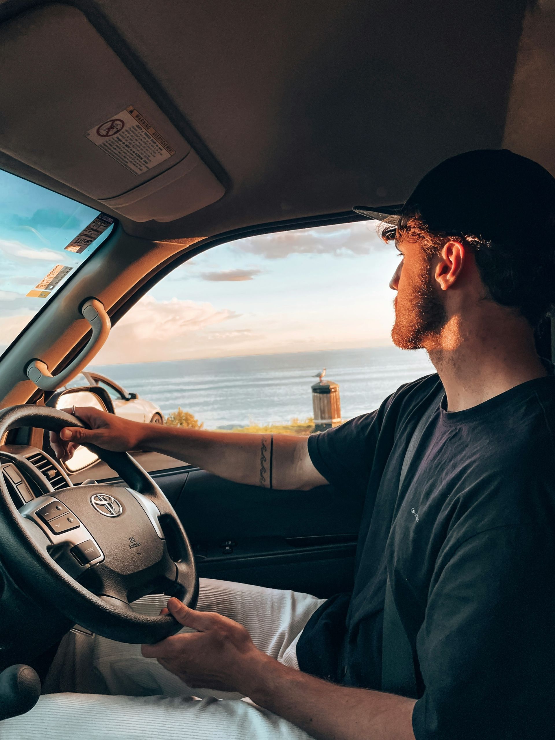 Man driving car, looking toward the ocean at sunset. Holds steering wheel.
