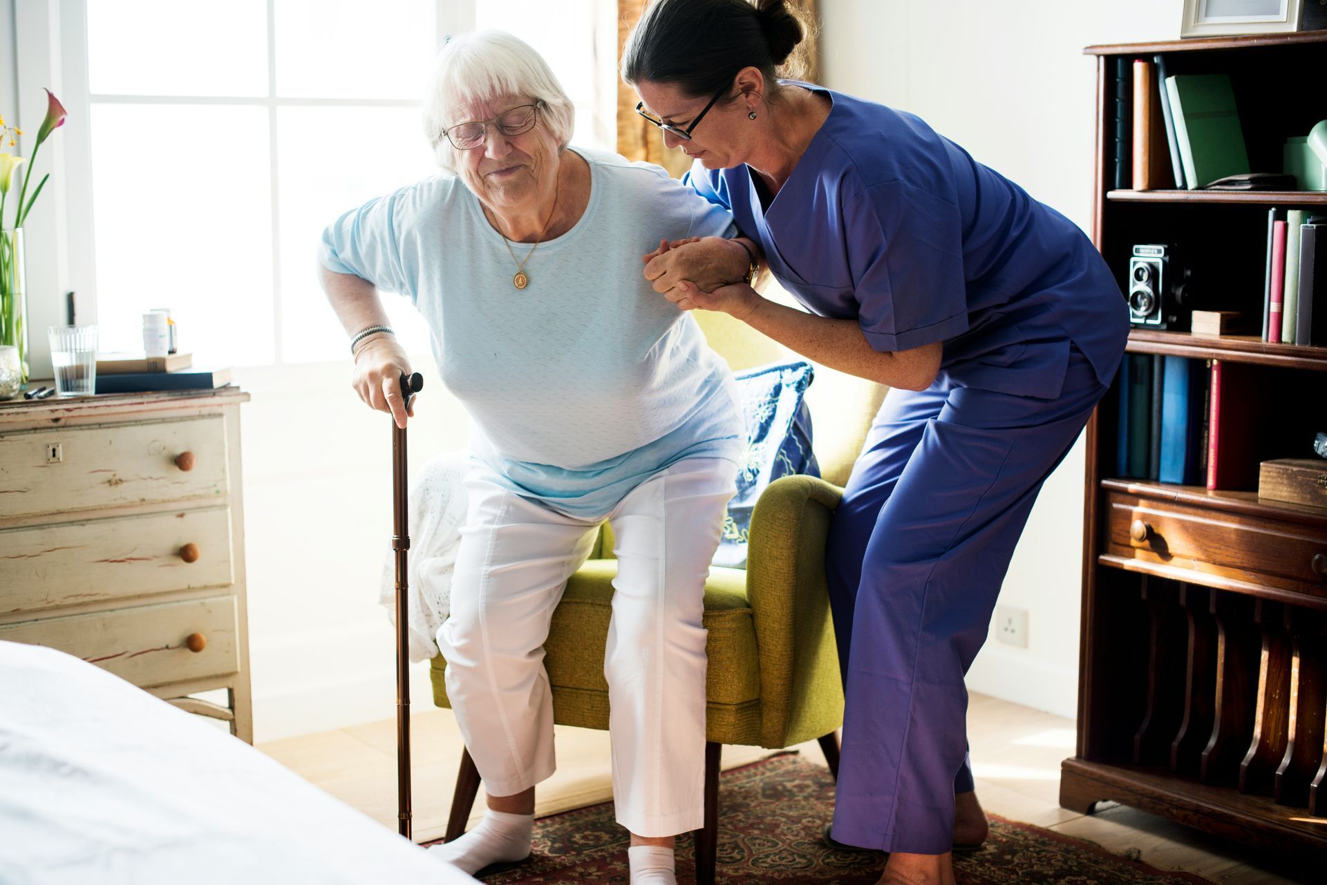 Caregiver assists an older person, using a cane, to stand up from a chair. Interior setting.
