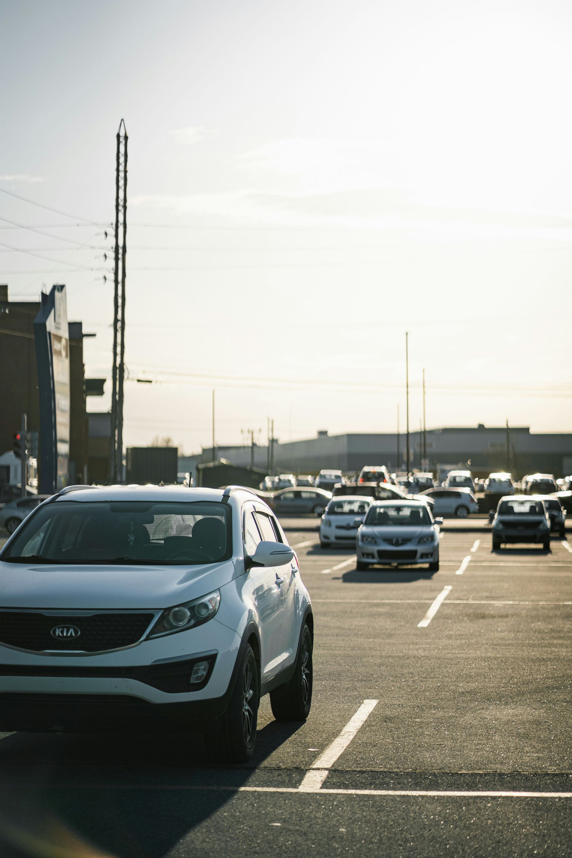 White Kia SUV parked in a lot, other cars visible in the background under a sunny sky.