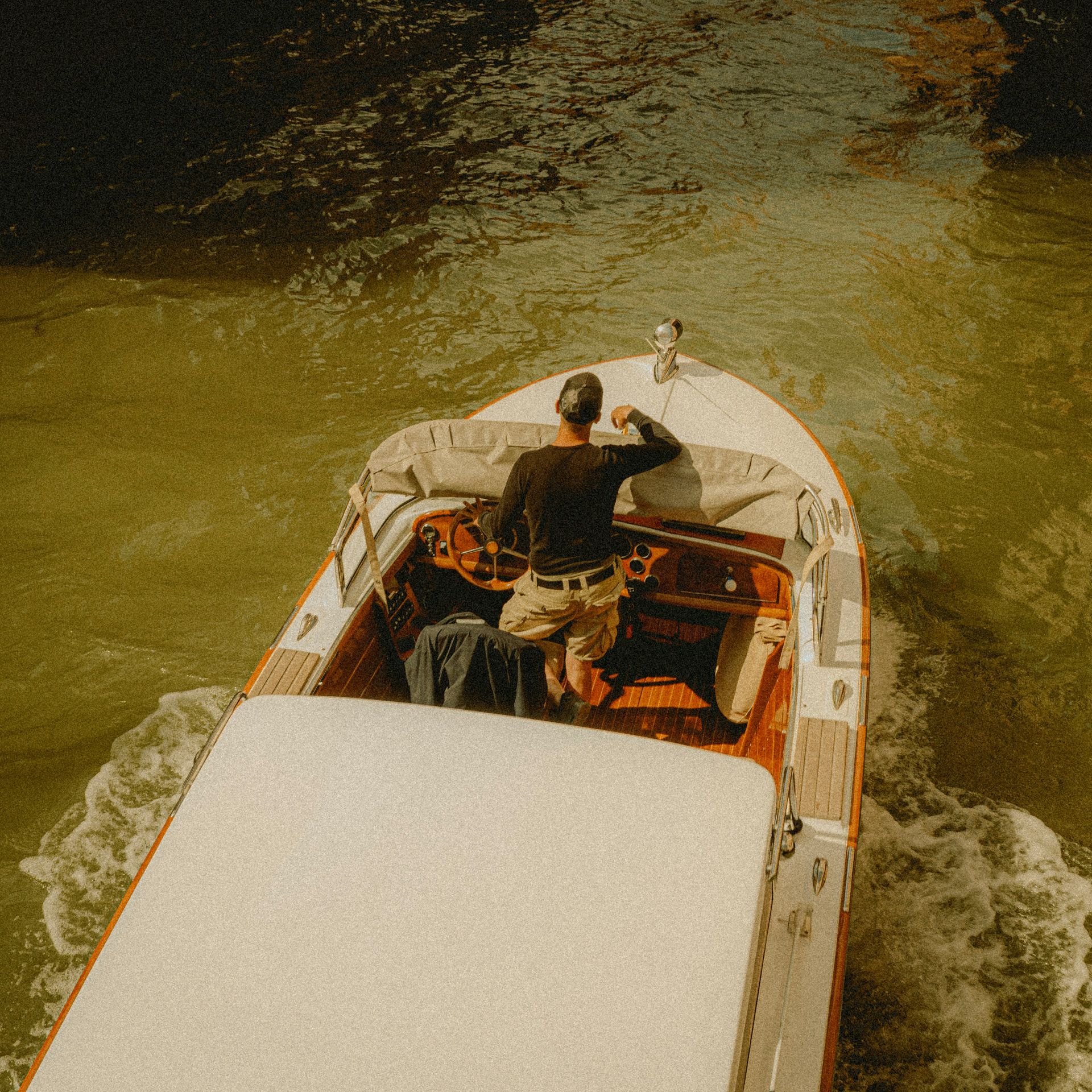 A man steers a wooden motorboat on a waterway, raising his arm, creating a wake.