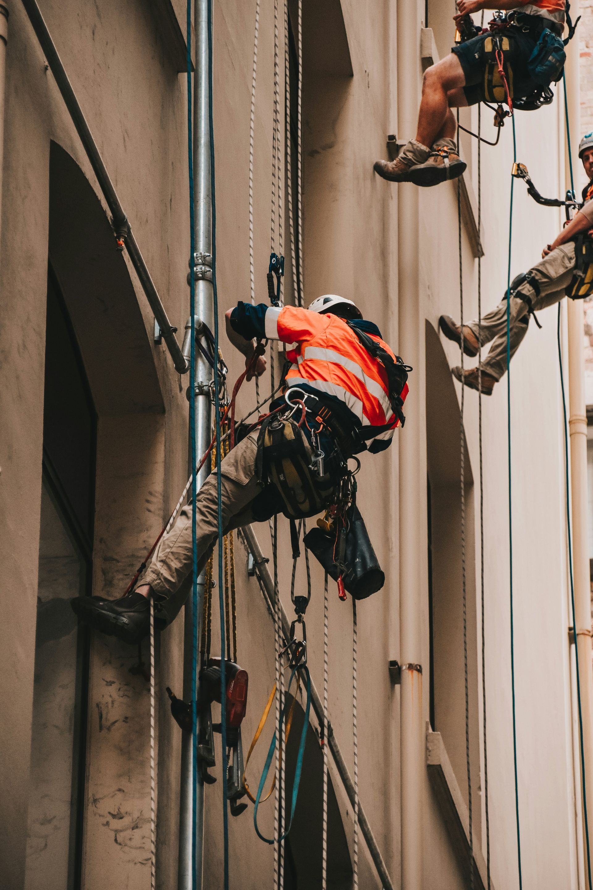 Workers in harnesses rappelling down a building facade, one wearing an orange safety vest.
