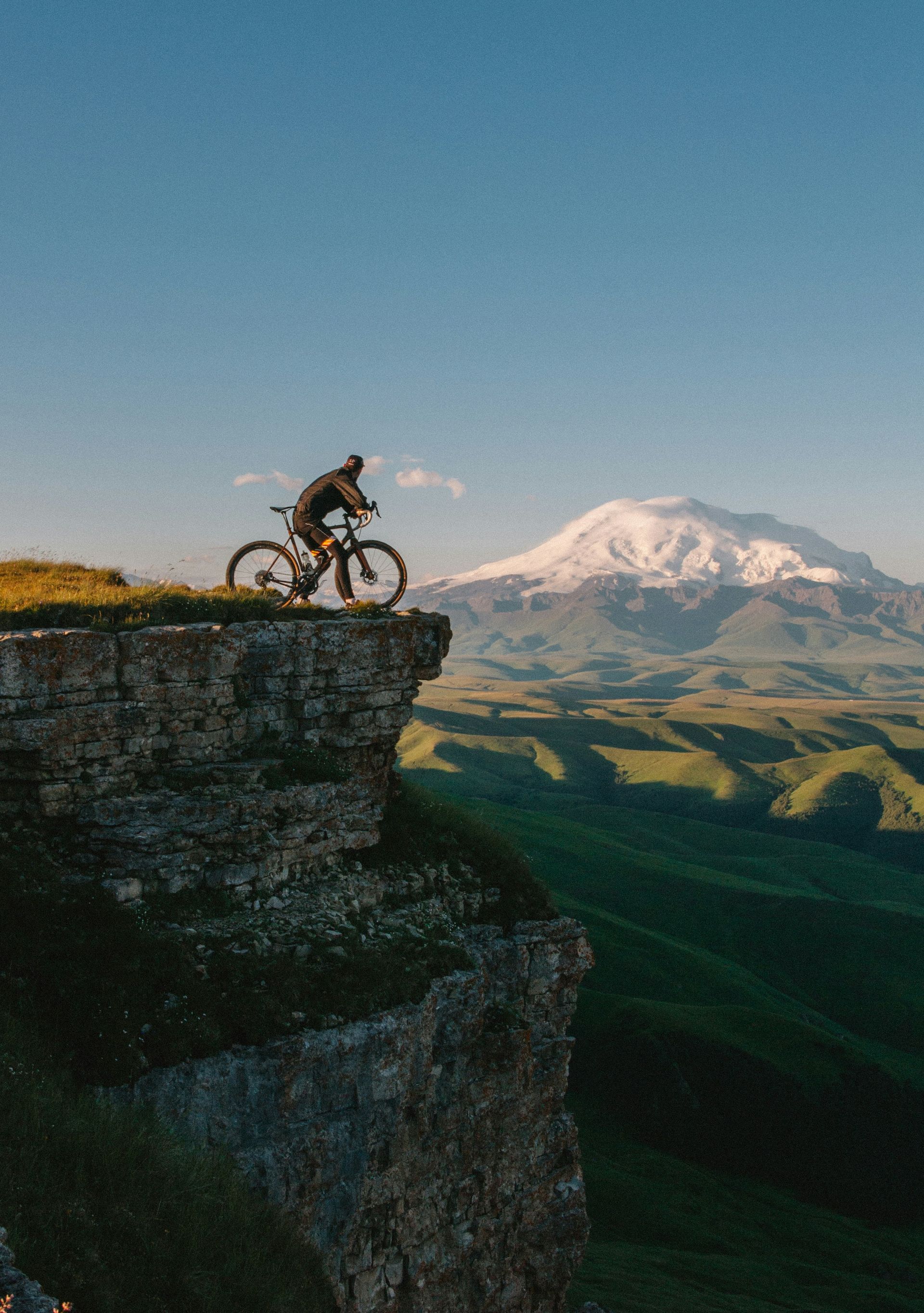 Mountain biker perched on a cliff edge, mountain landscape in background, blue sky.