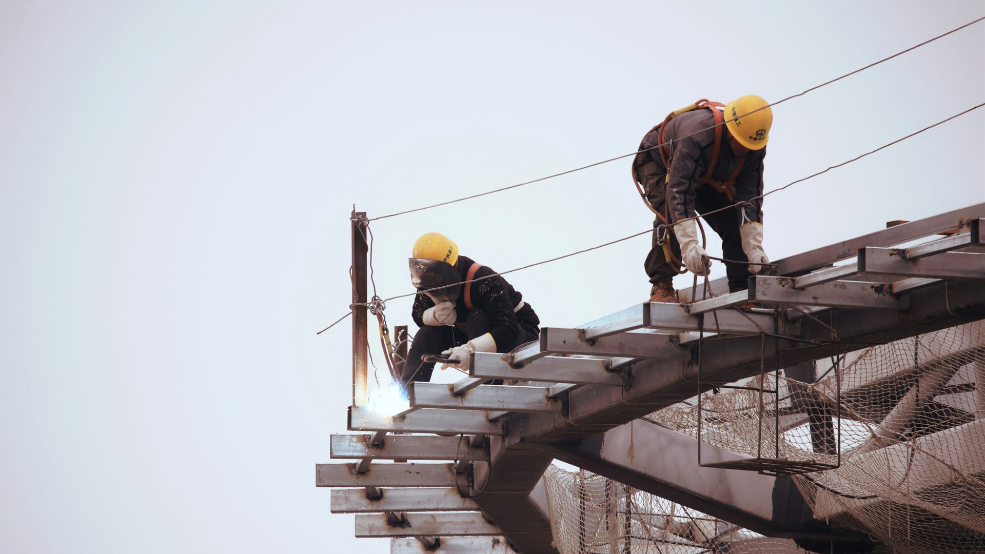 Two construction workers welding metal beams, wearing hard hats, outdoors.
