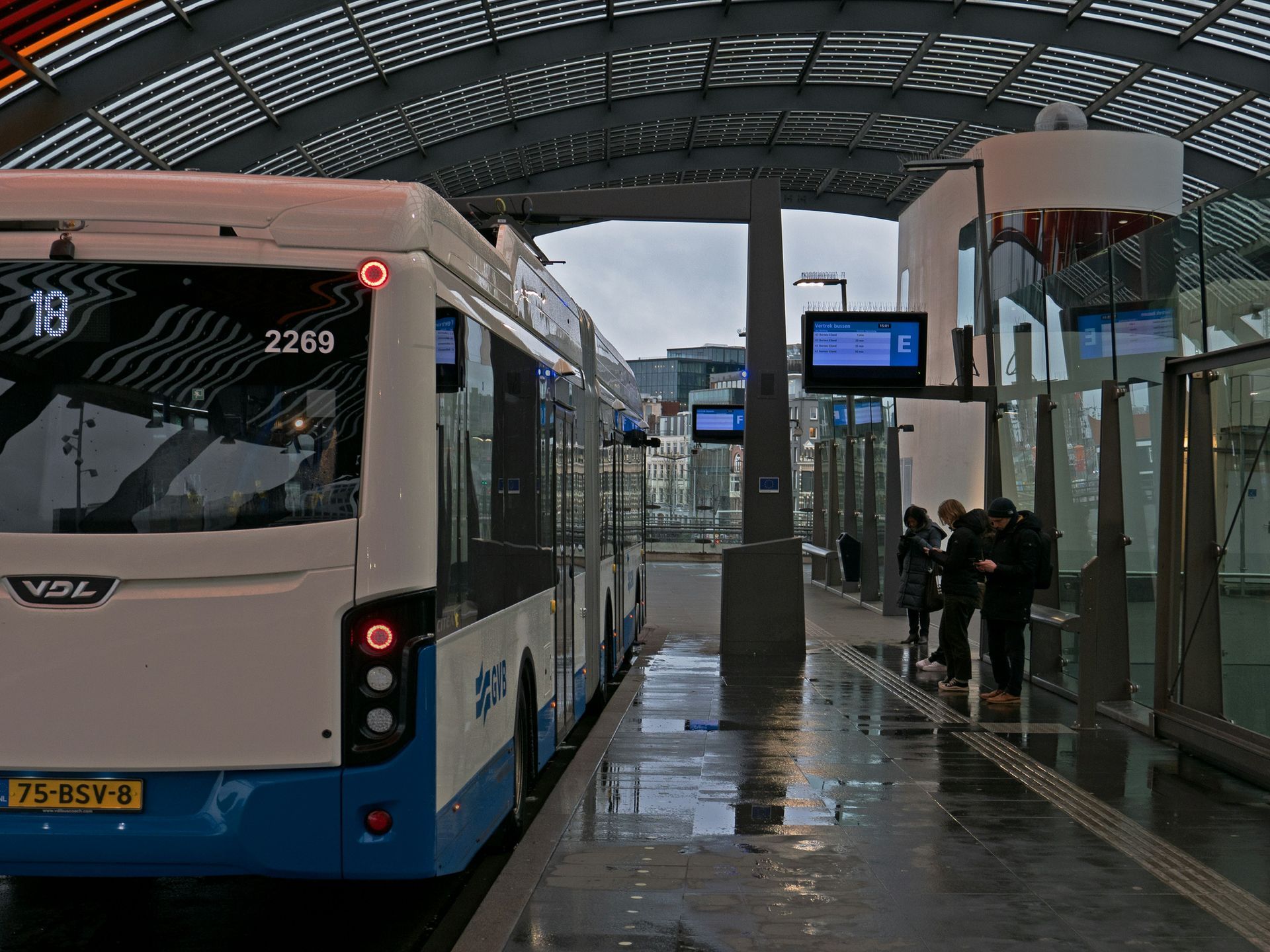 Bus at a covered bus station, passengers waiting. White and blue bus, gray structure.