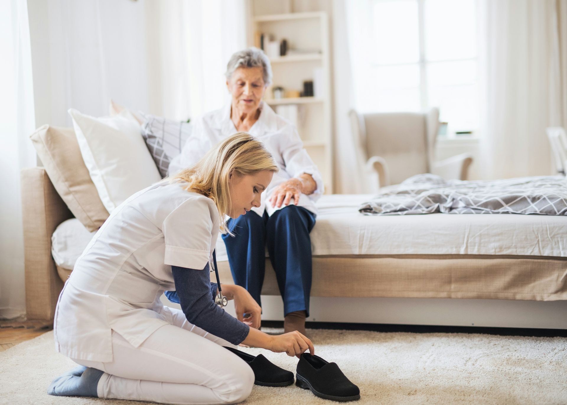 Nurse kneeling, putting shoes on person sitting on bed in bedroom.
