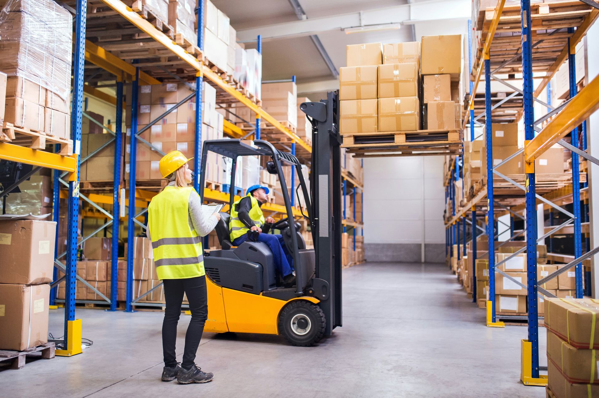 Warehouse scene: Worker in yellow safety vest directs forklift driver amidst stacked boxes.