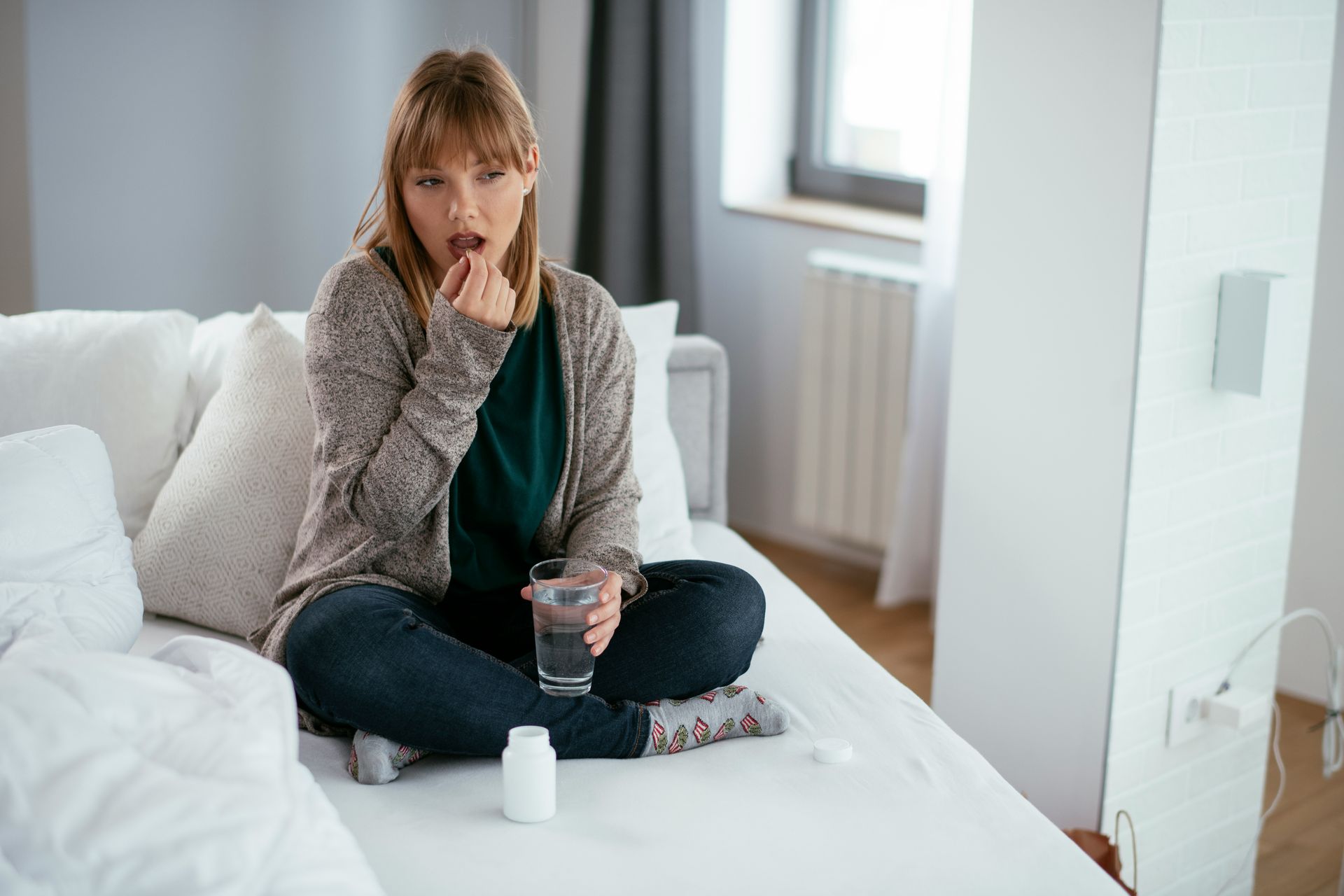 Woman on bed, taking a pill. Holds a glass of water, bottle of pills nearby. Indoors.