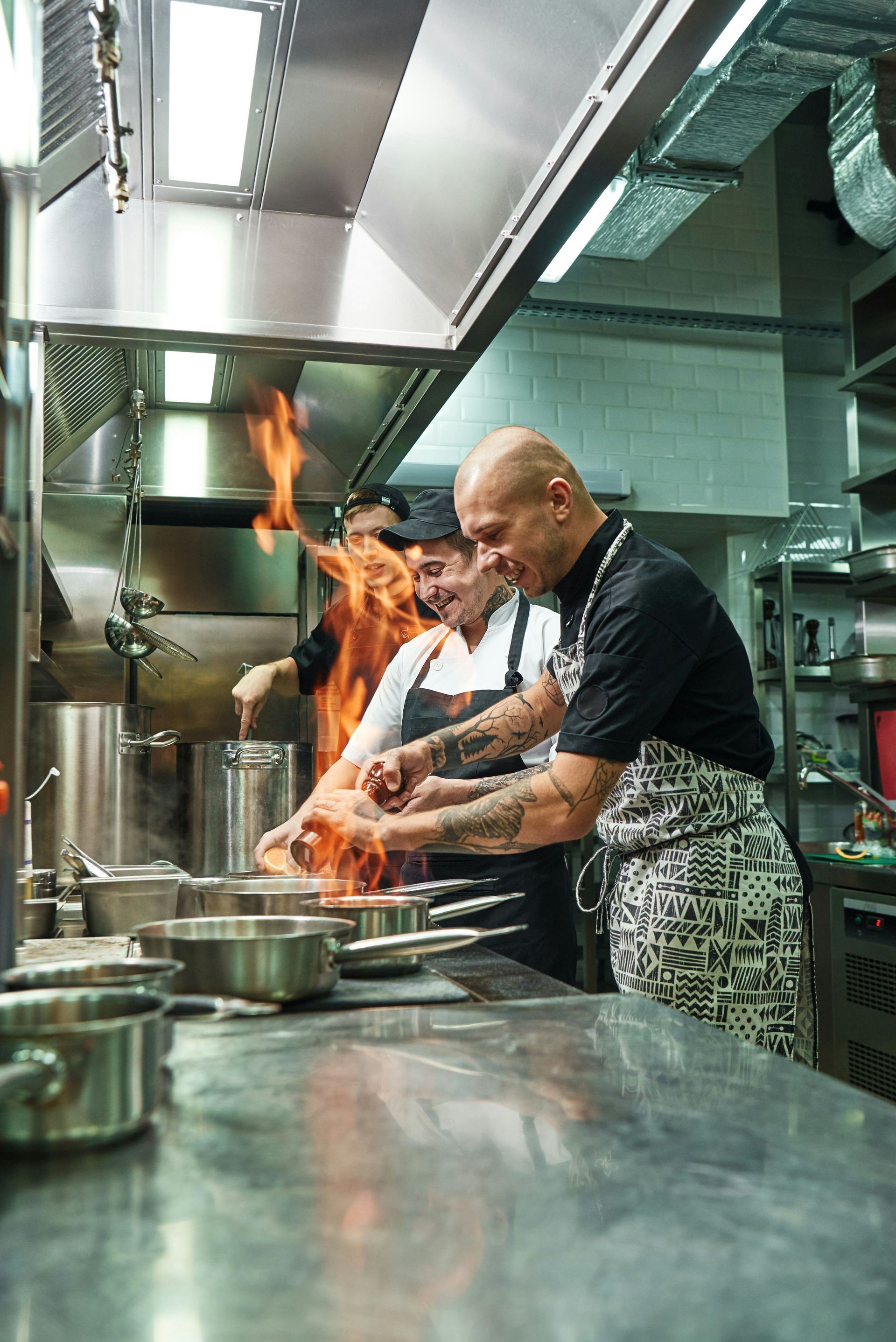 Chefs cooking in a commercial kitchen with flames; stainless steel surfaces.