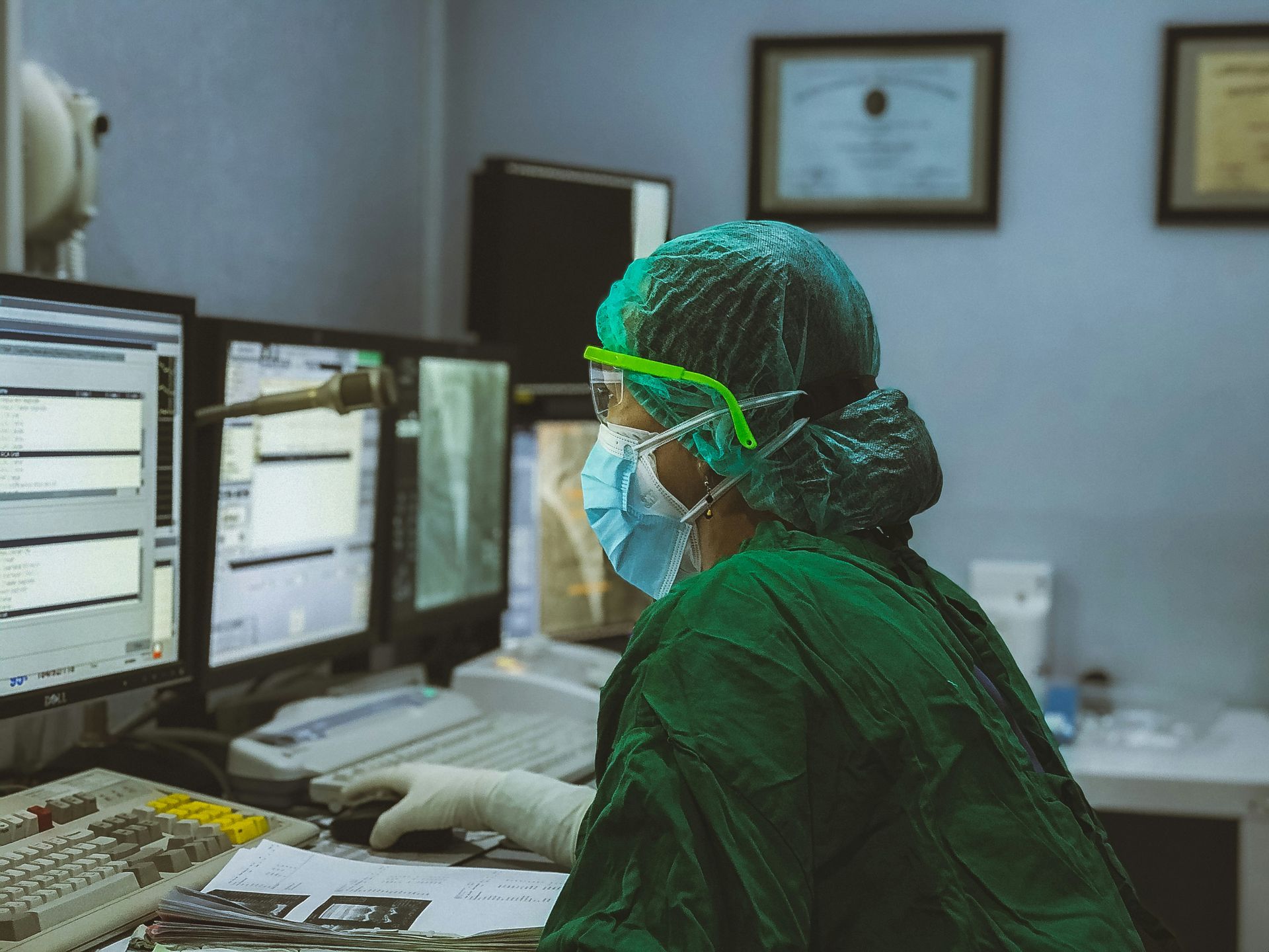 Medical professional in scrubs and mask operating computer monitors in a medical setting.