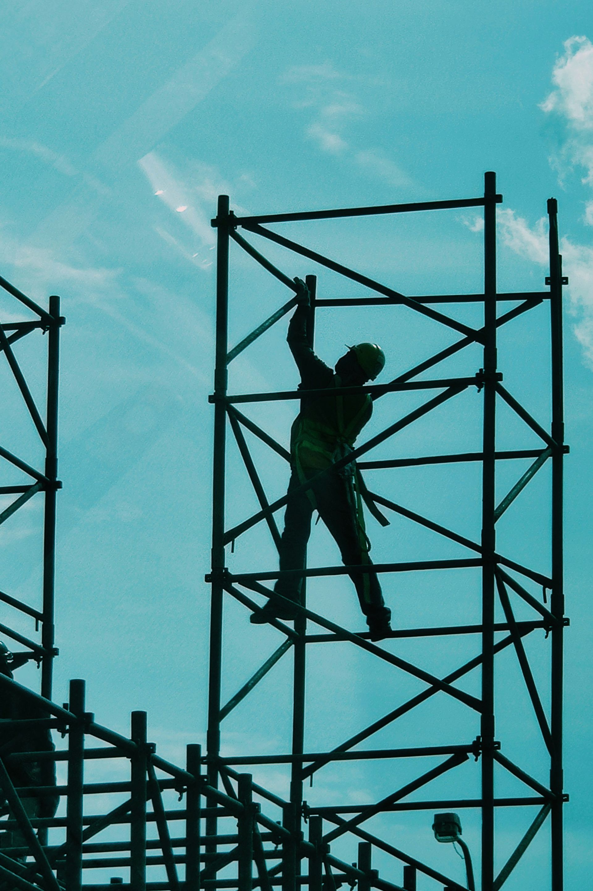 Construction worker climbing a metal scaffold against a teal-toned sky.
