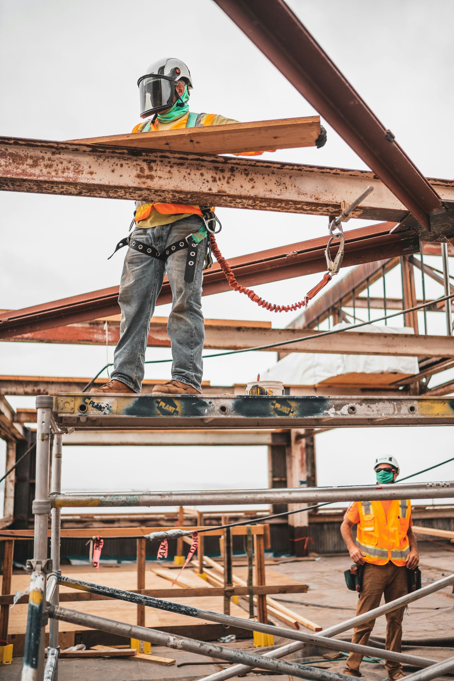 Two construction workers on scaffolding. One in safety gear holds wood beam, the other stands below.