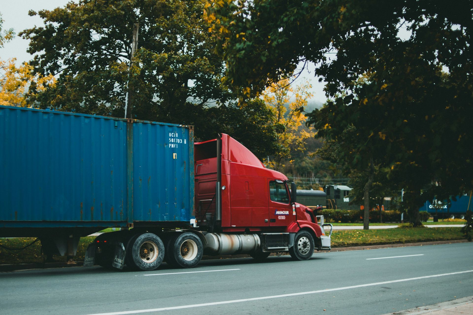 Red semi-truck with blue container parked on the side of a road, trees in the background.