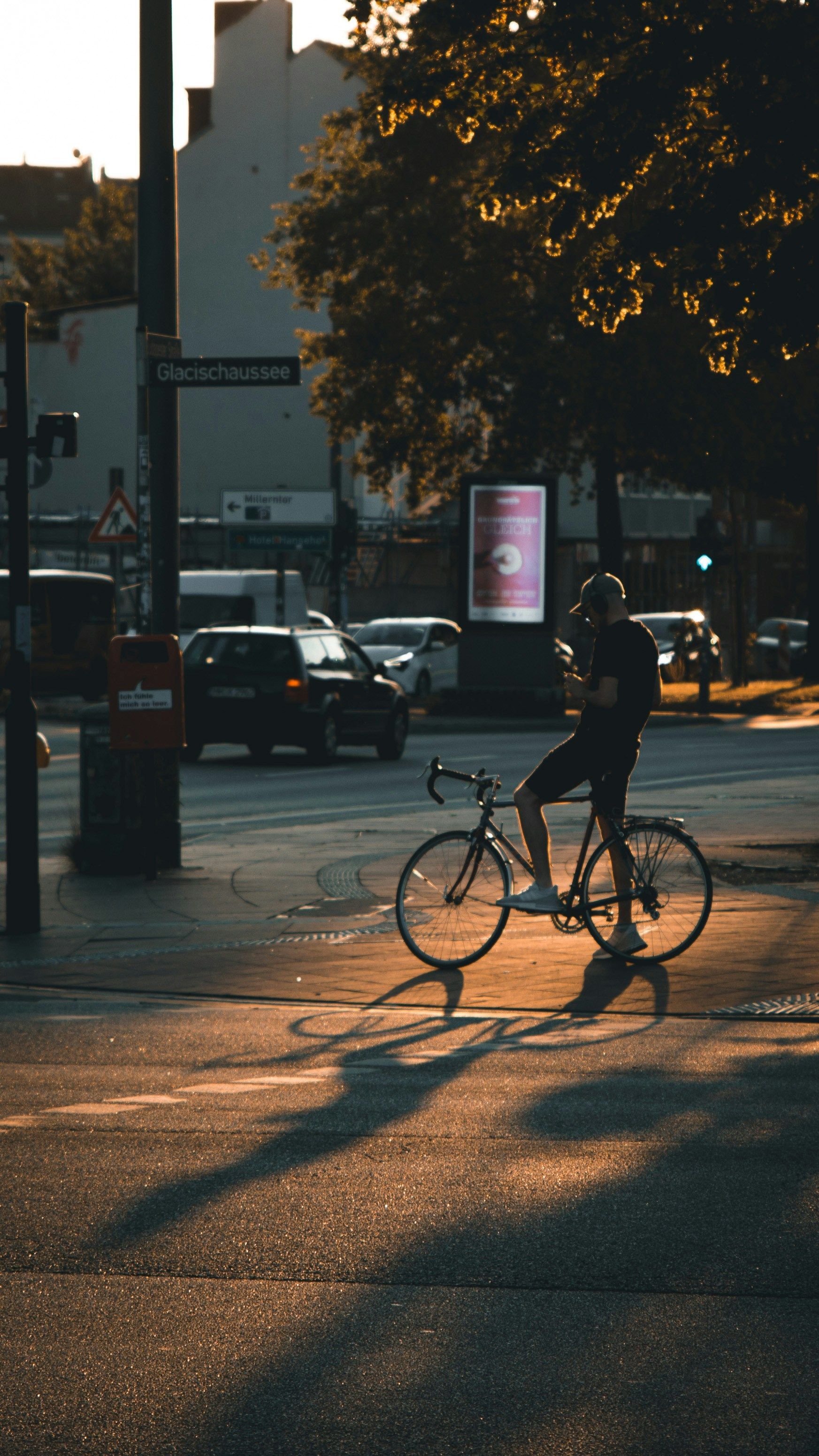 Cyclist on bike at a crosswalk, waiting for traffic light. Golden hour, city scene with cars and buildings.