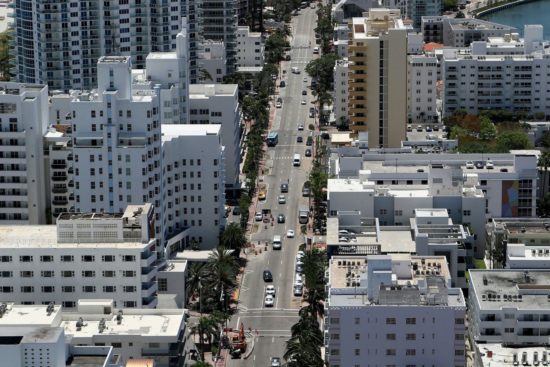 Aerial view of a city street lined with white buildings and palm trees, with cars driving down the road.
