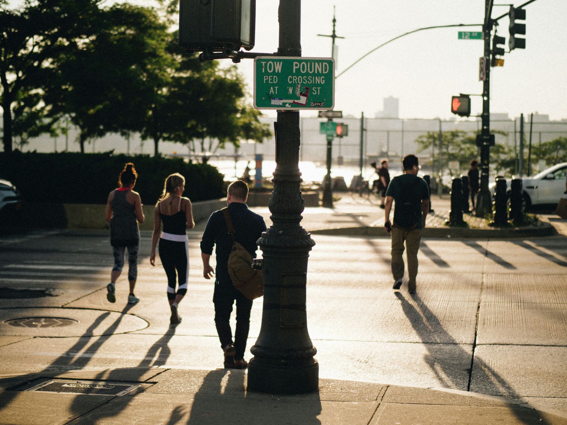 People walking on a city crosswalk in the sunlight, past a street sign and a black lamp post.