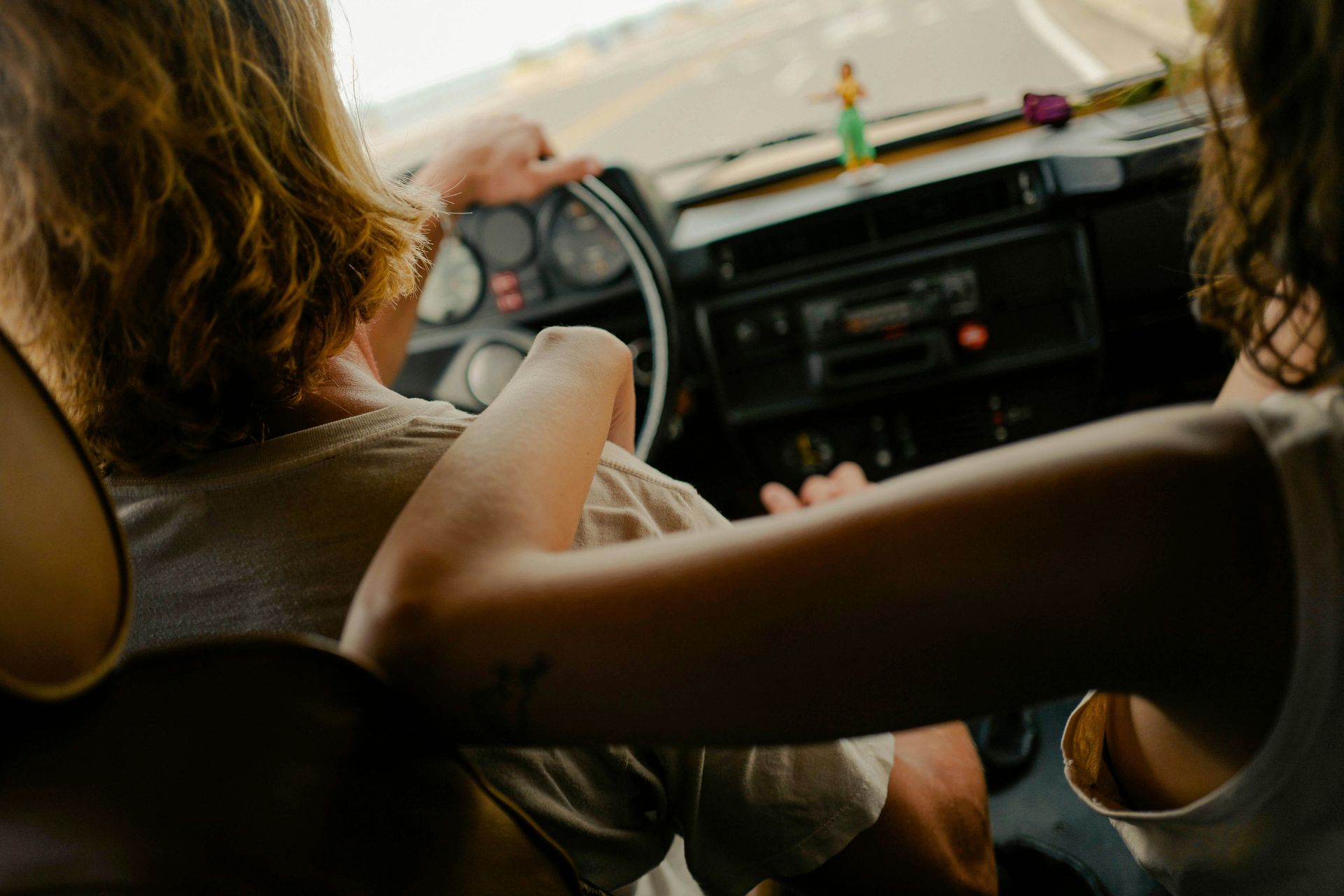Two people in a vehicle, one driving. One person's arm rests on the other's shoulder. Interior view with dashboard.