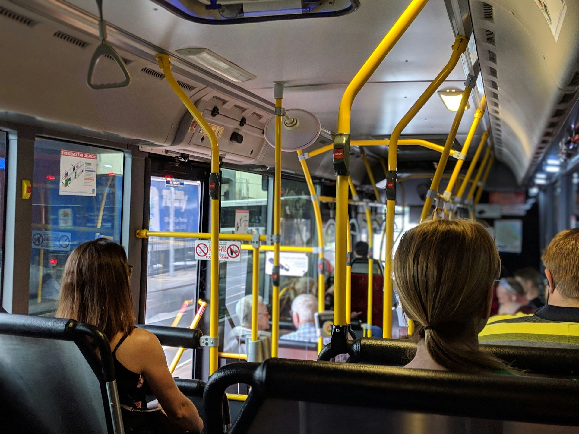 Interior view of a bus with passengers seated. Yellow handrails and window reflections visible.