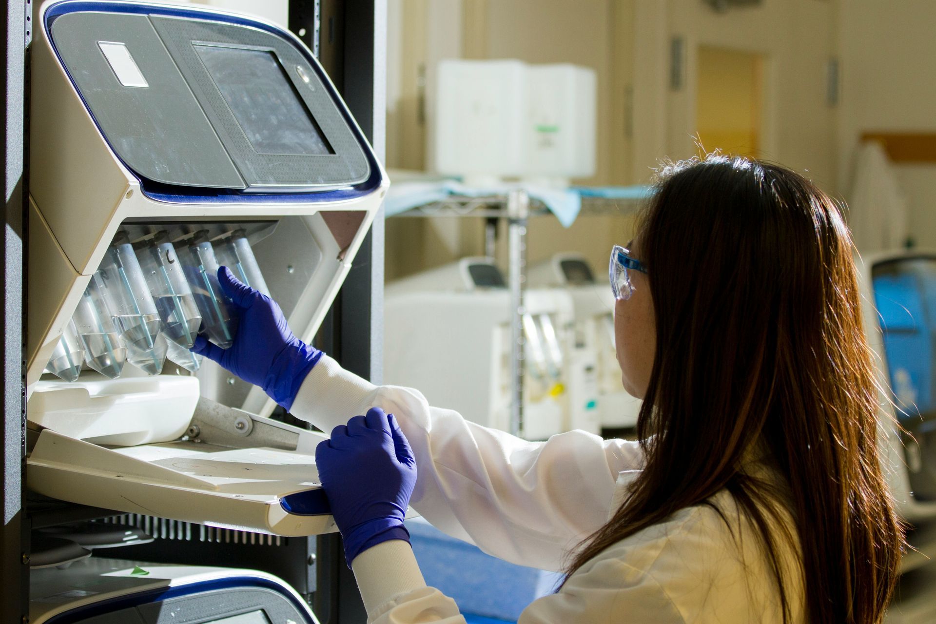Woman in lab coat and gloves placing vials into a scientific instrument.