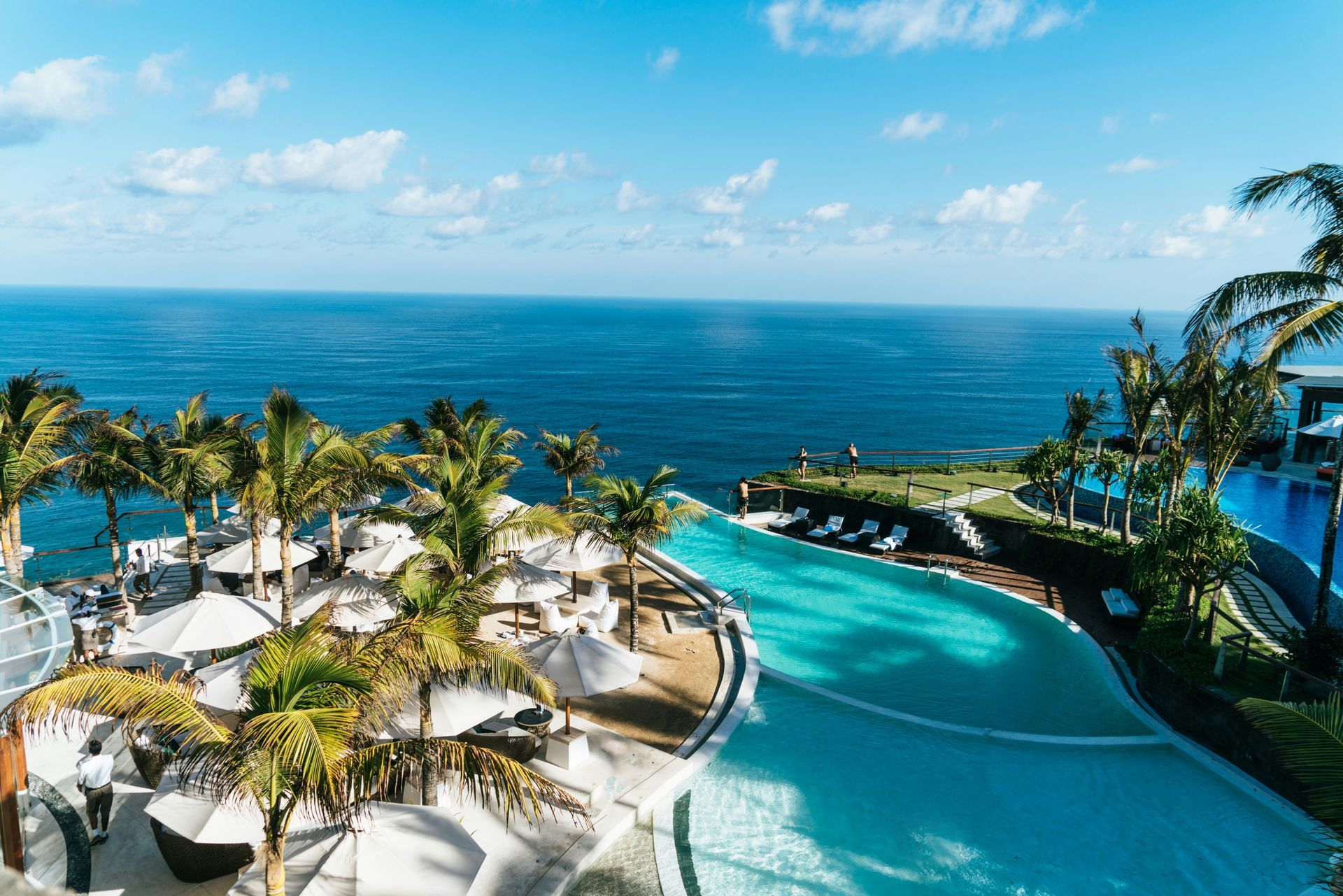 Swimming pool overlooking ocean; palm trees, white umbrellas, blue sky.