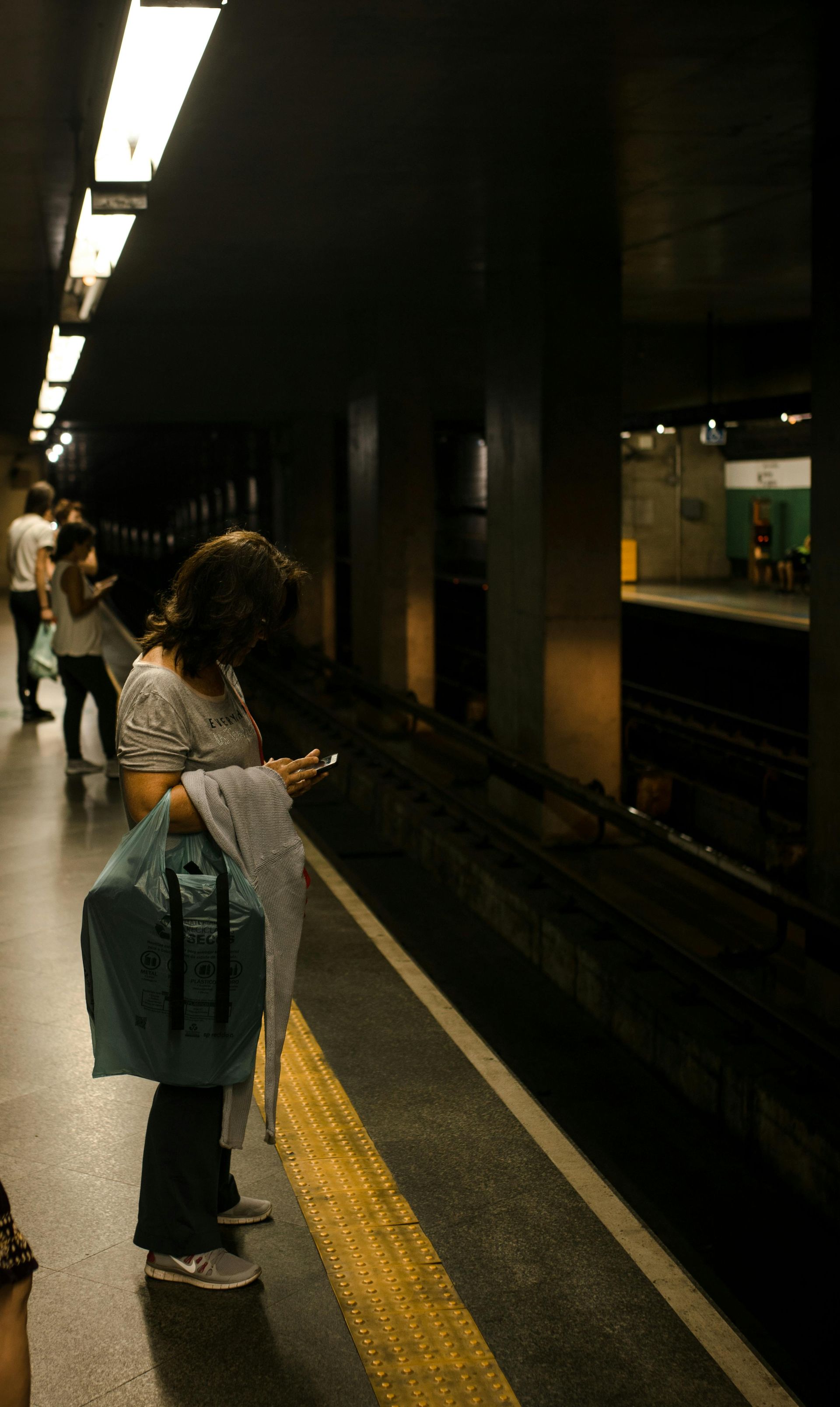 People waiting on a subway platform. Woman checks her phone, dark setting with overhead lights.