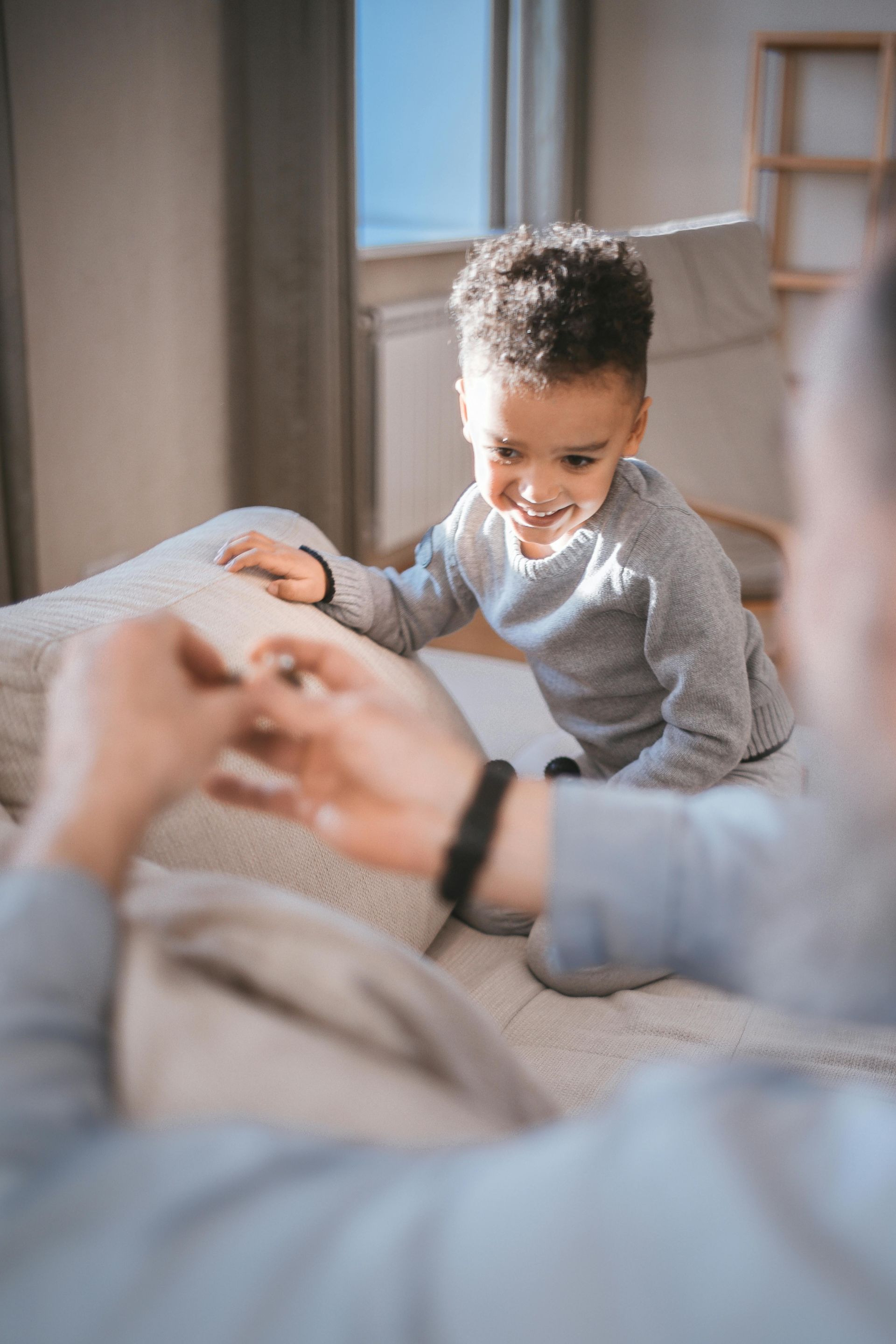 Child on a bed smiles at a person, reaching out. Bright room with window in background.