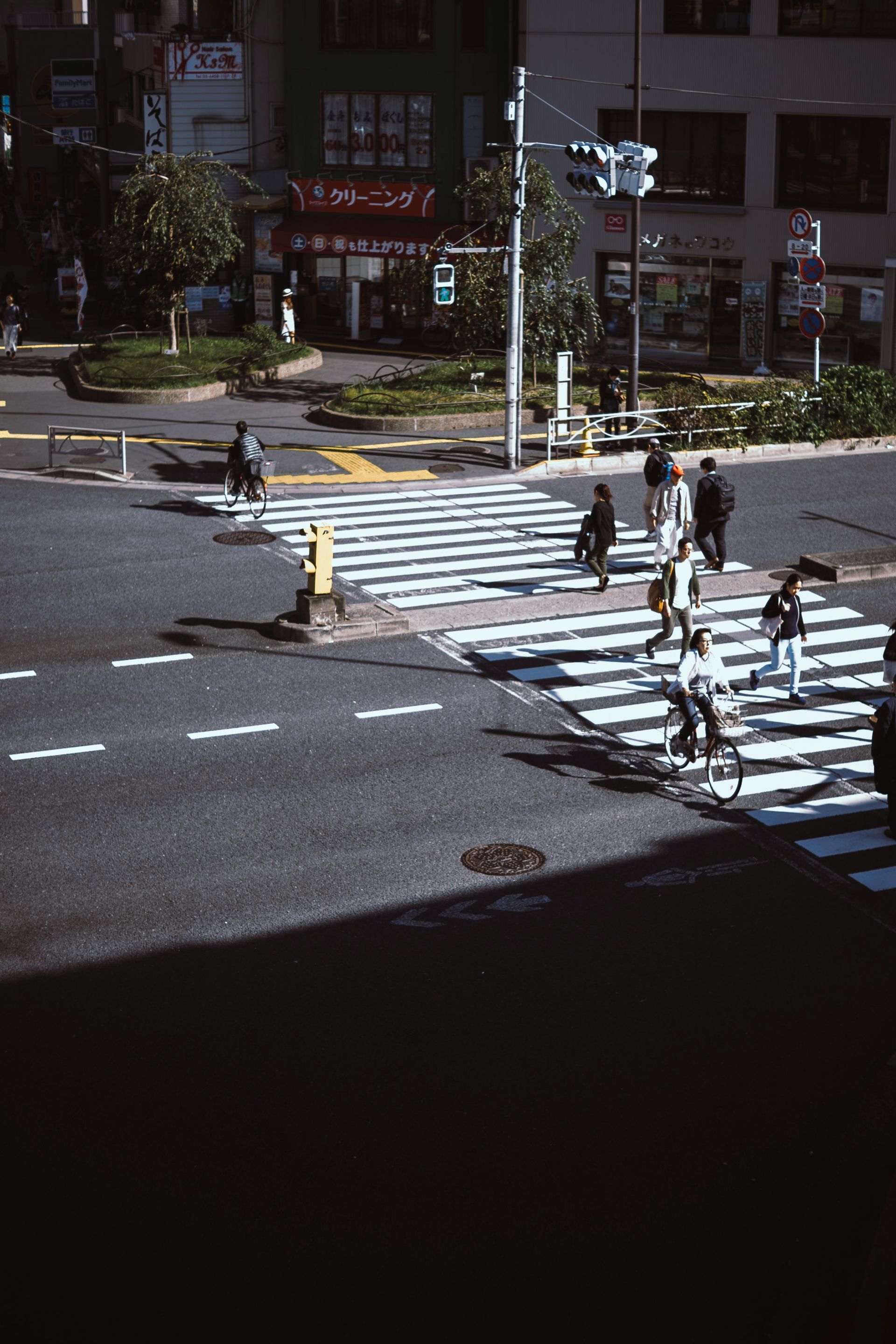 Busy city intersection with pedestrians crossing a crosswalk and a cyclist. Buildings and a traffic light are visible.