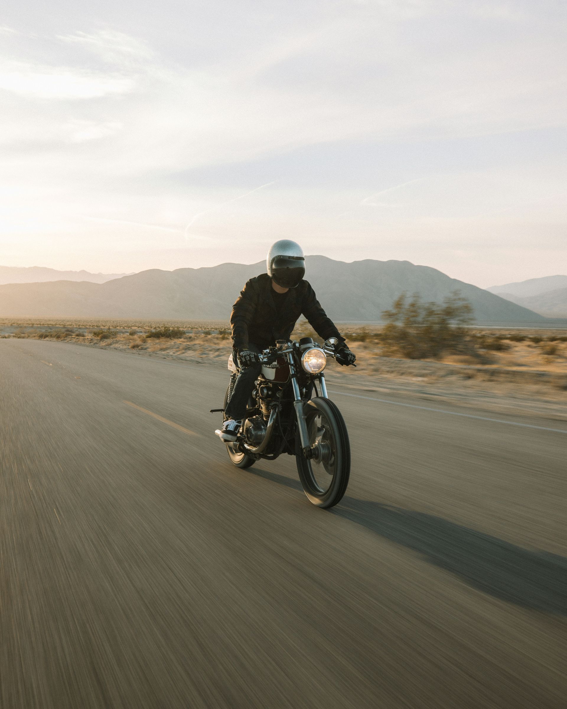 Motorcyclist riding on a desert road, wearing a helmet and dark clothing; mountains in the background.