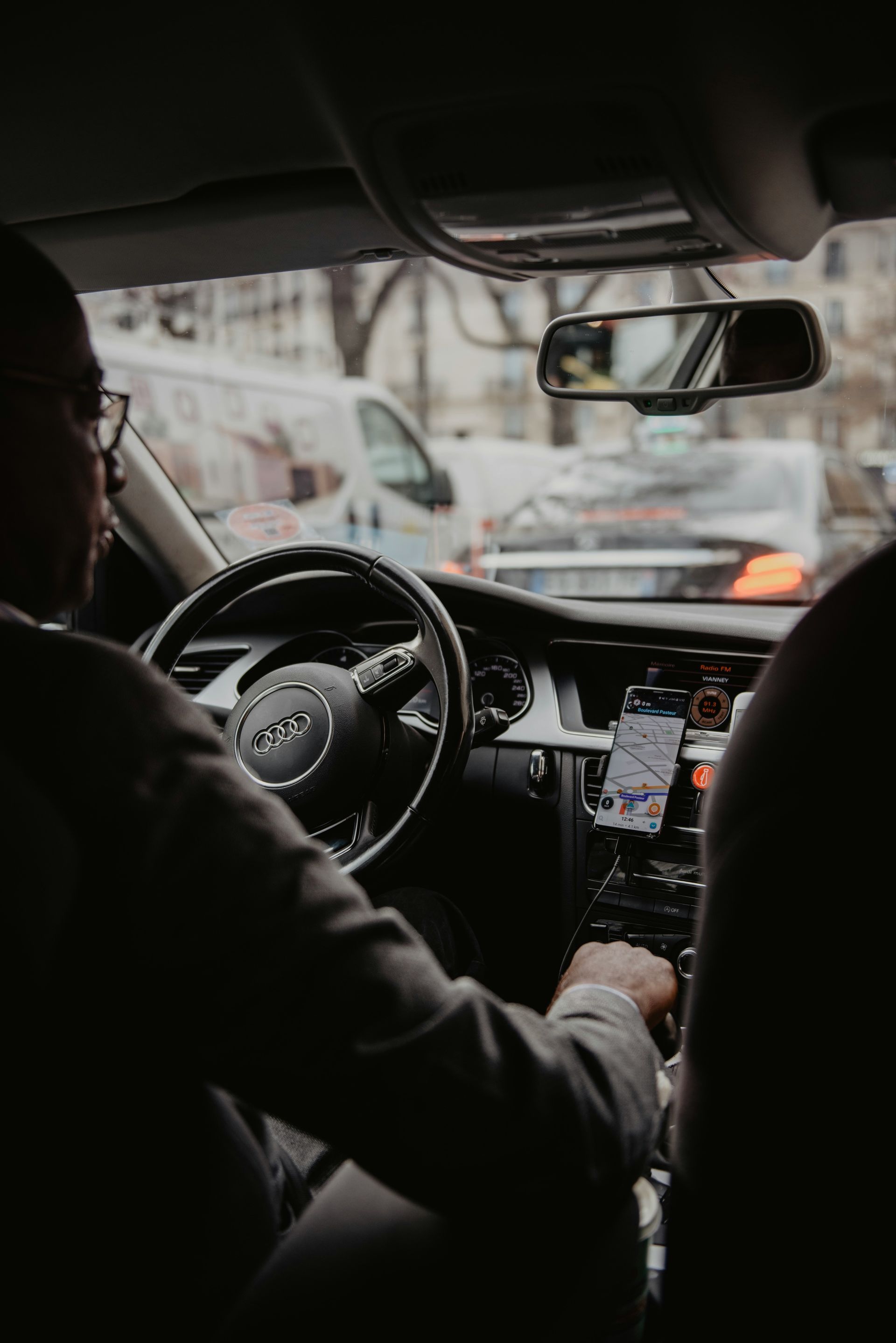 Person driving an Audi, seen from the backseat, looking ahead. Dashboard visible, phone mounted, and street scene in the background.