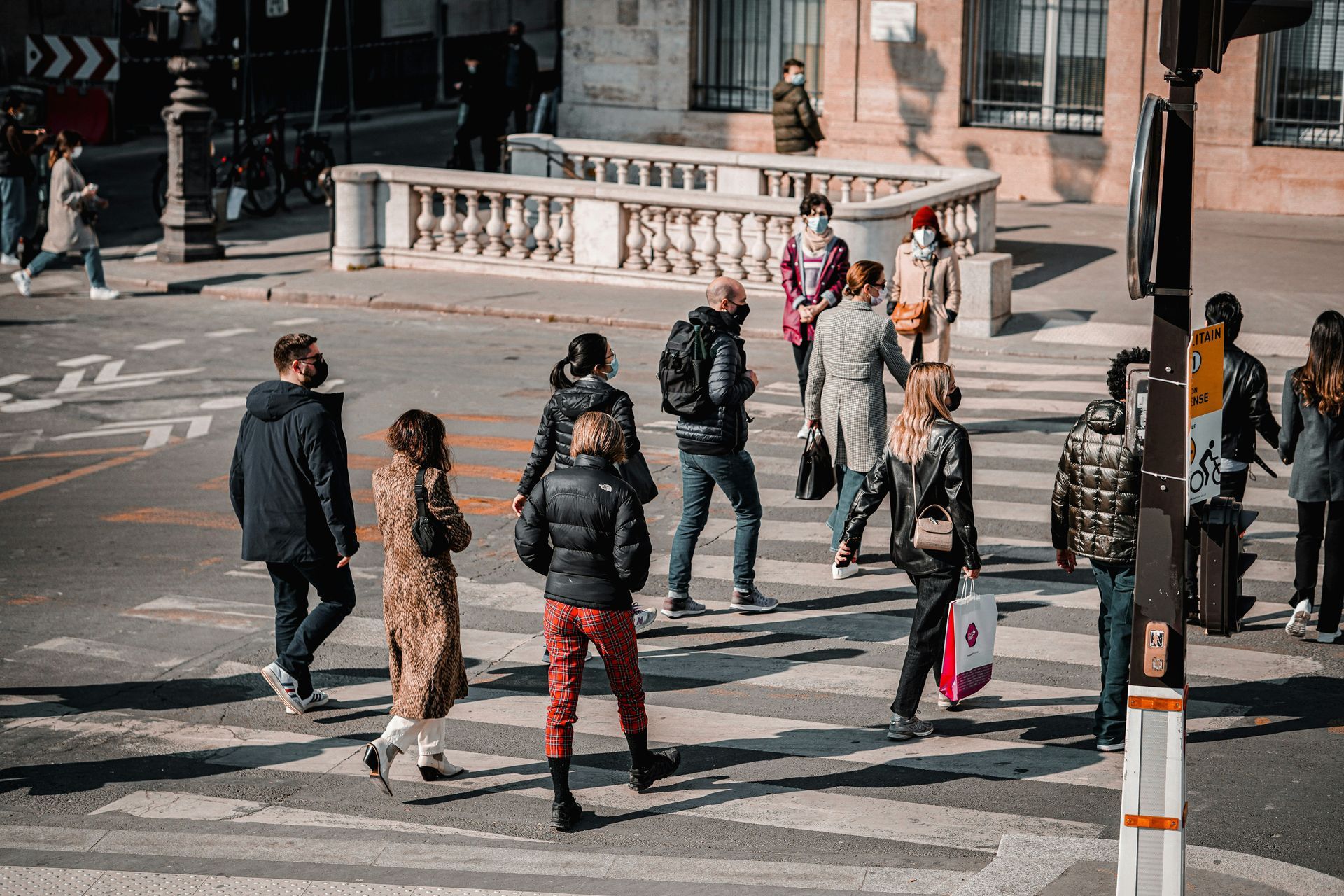 People wearing masks crossing a city street at a crosswalk with buildings in the background.