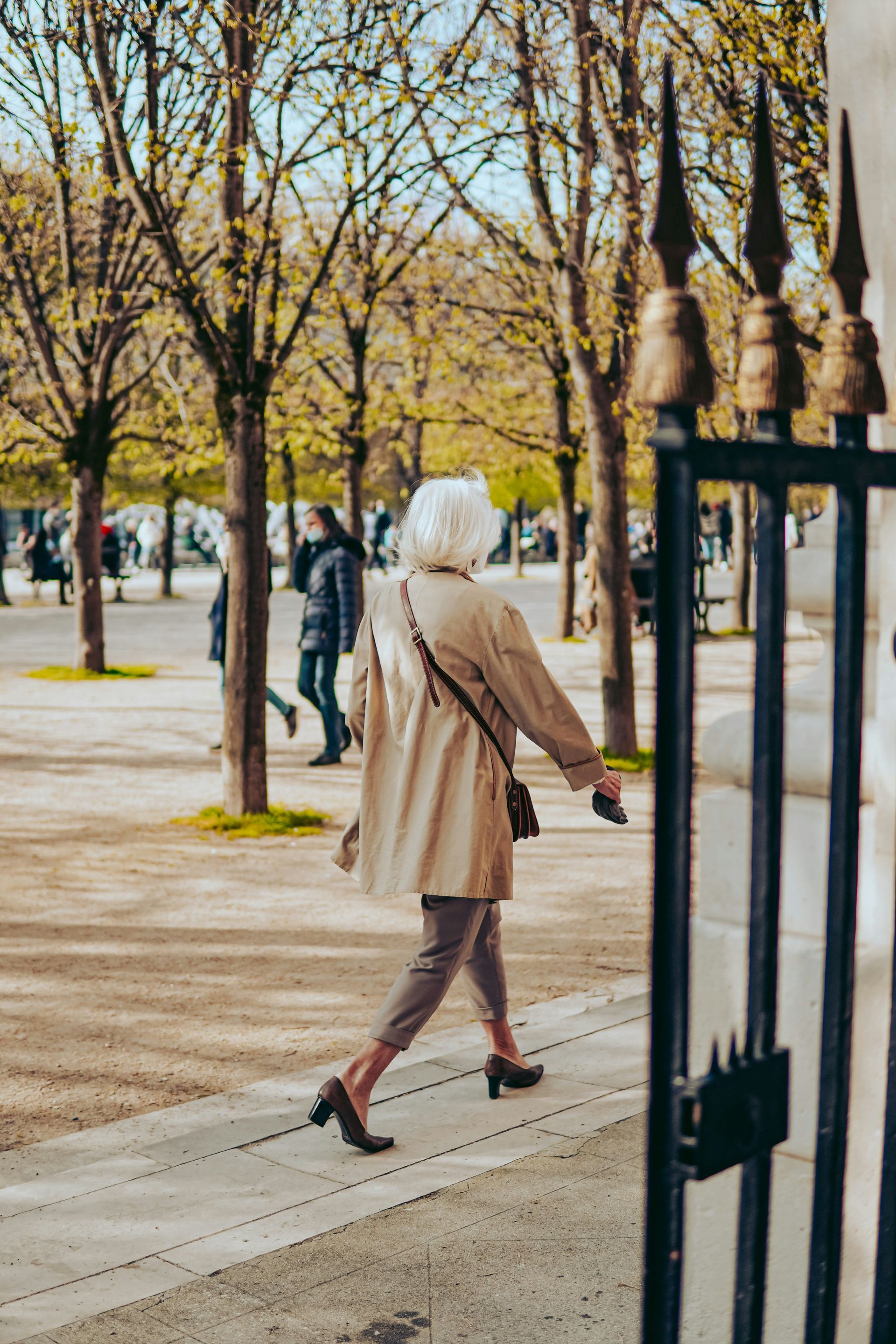 Woman in beige coat and heels walks along a path past a wrought-iron fence; park setting.