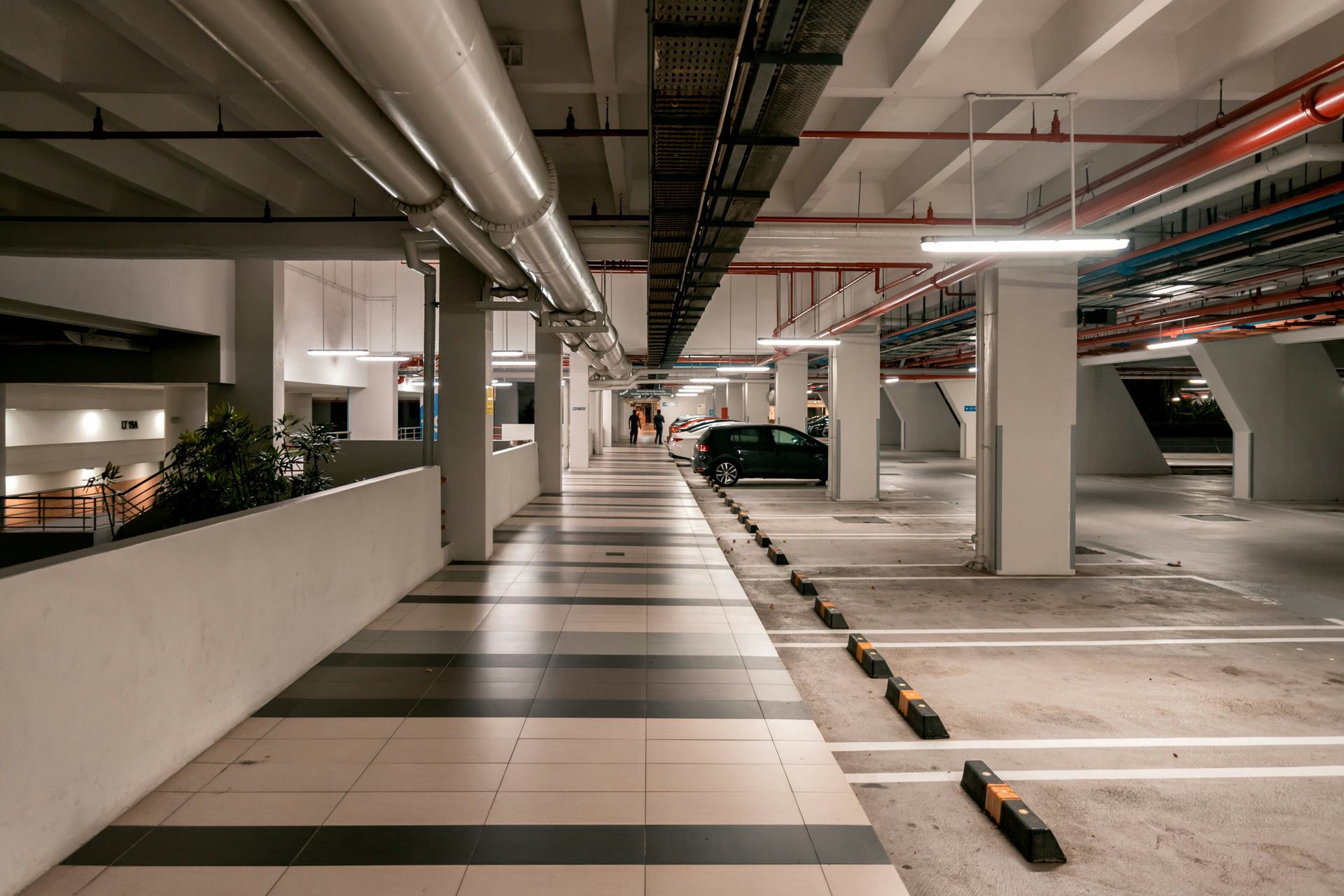 Underground parking garage with columns, pipes, and parked car. Striped floor, people walking in the distance.
