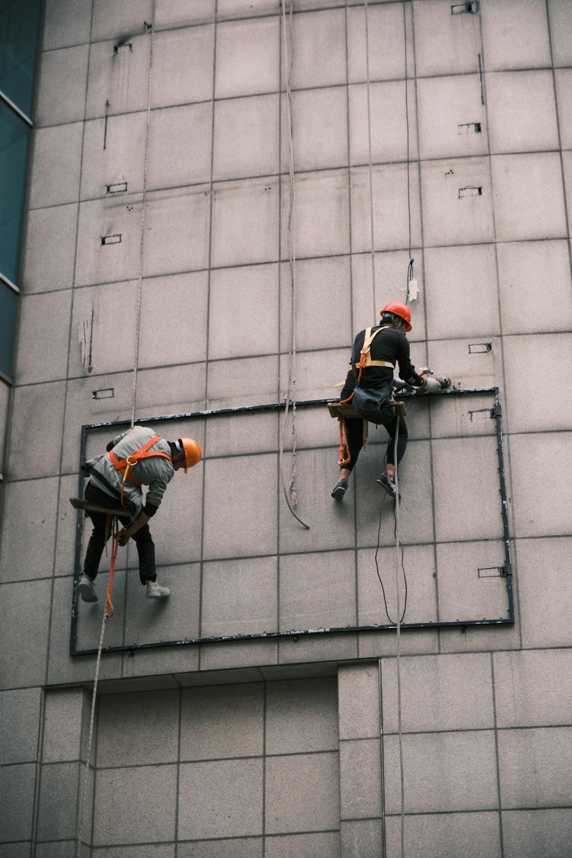Two workers in safety gear cleaning the facade of a building, using ropes to descend and tools to work on the wall.