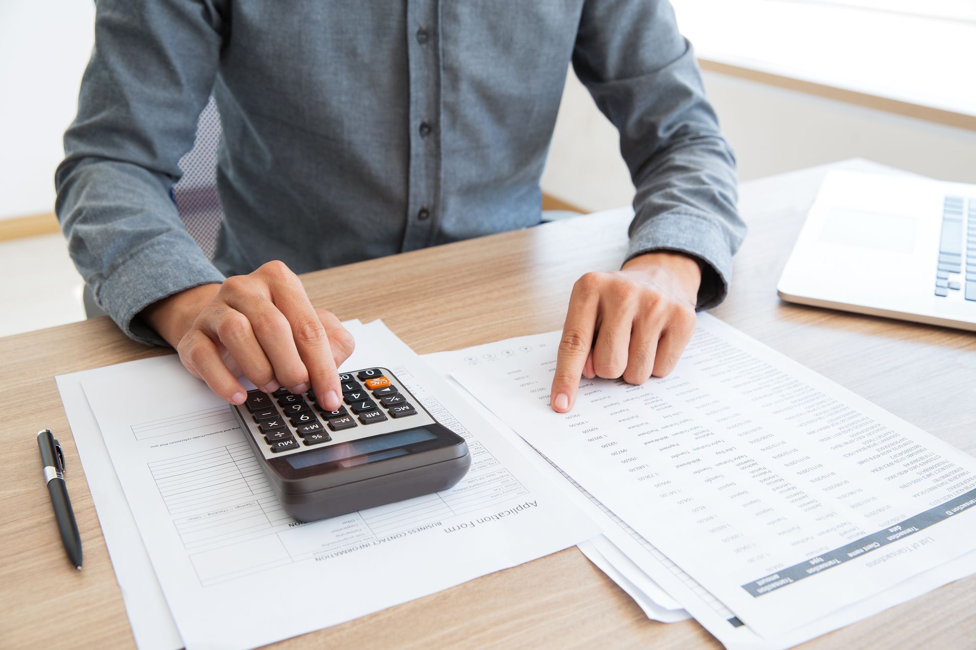 Person in a white shirt using a calculator at a wooden desk, writing and reviewing financial data.