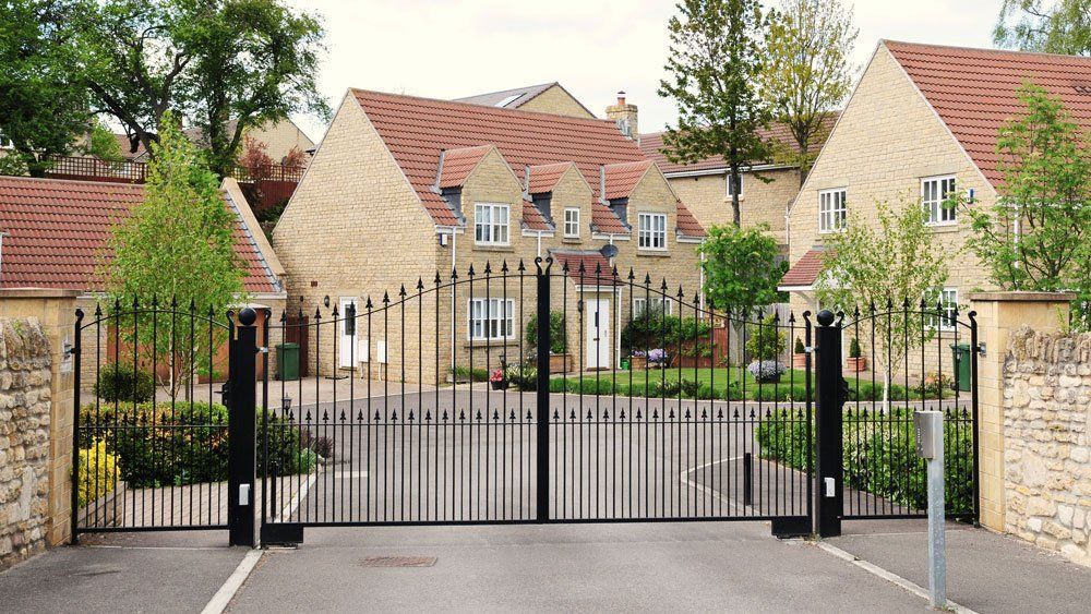 Gated entrance to a modern house with stone walls, black iron gate, and red-tiled roofs behind greenery
