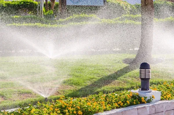 Lawn sprinkler spraying water over a flower bed and grass near a lamp post and tree trunk