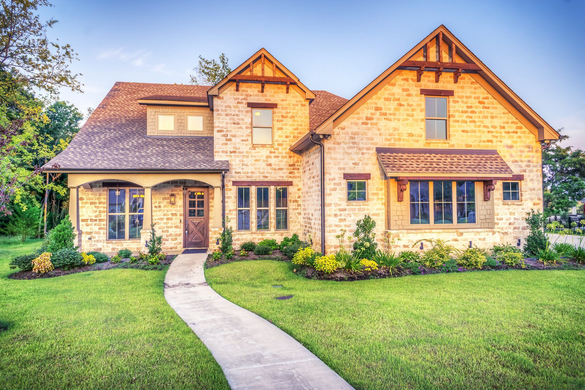 Stone house with tan roof, front walkway, and manicured green lawn in a sunny suburban setting