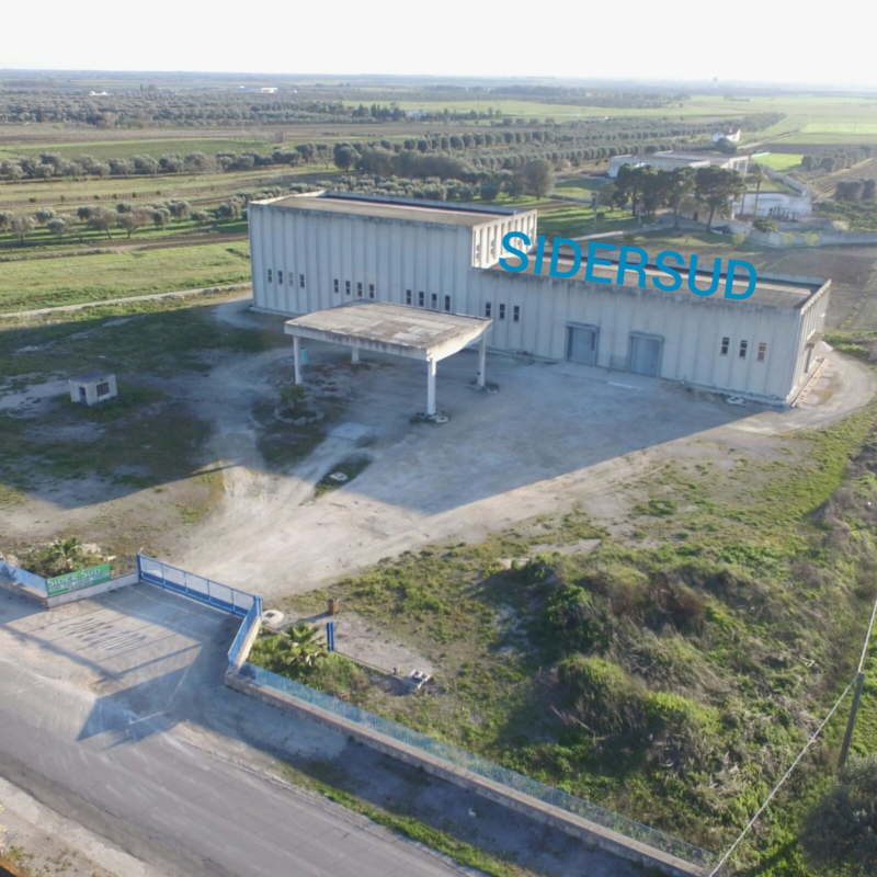 Veduta aerea di un grande edificio industriale in cemento con un'insegna blu, circondato da campi aperti e strade.