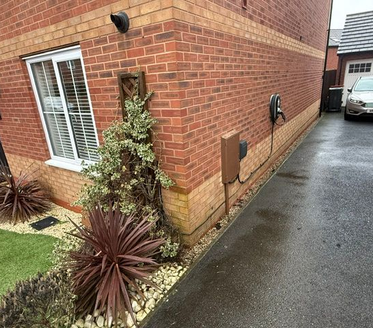 Brick house corner with narrow driveway, wet pavement, and a small planted garden bed with shrubs.