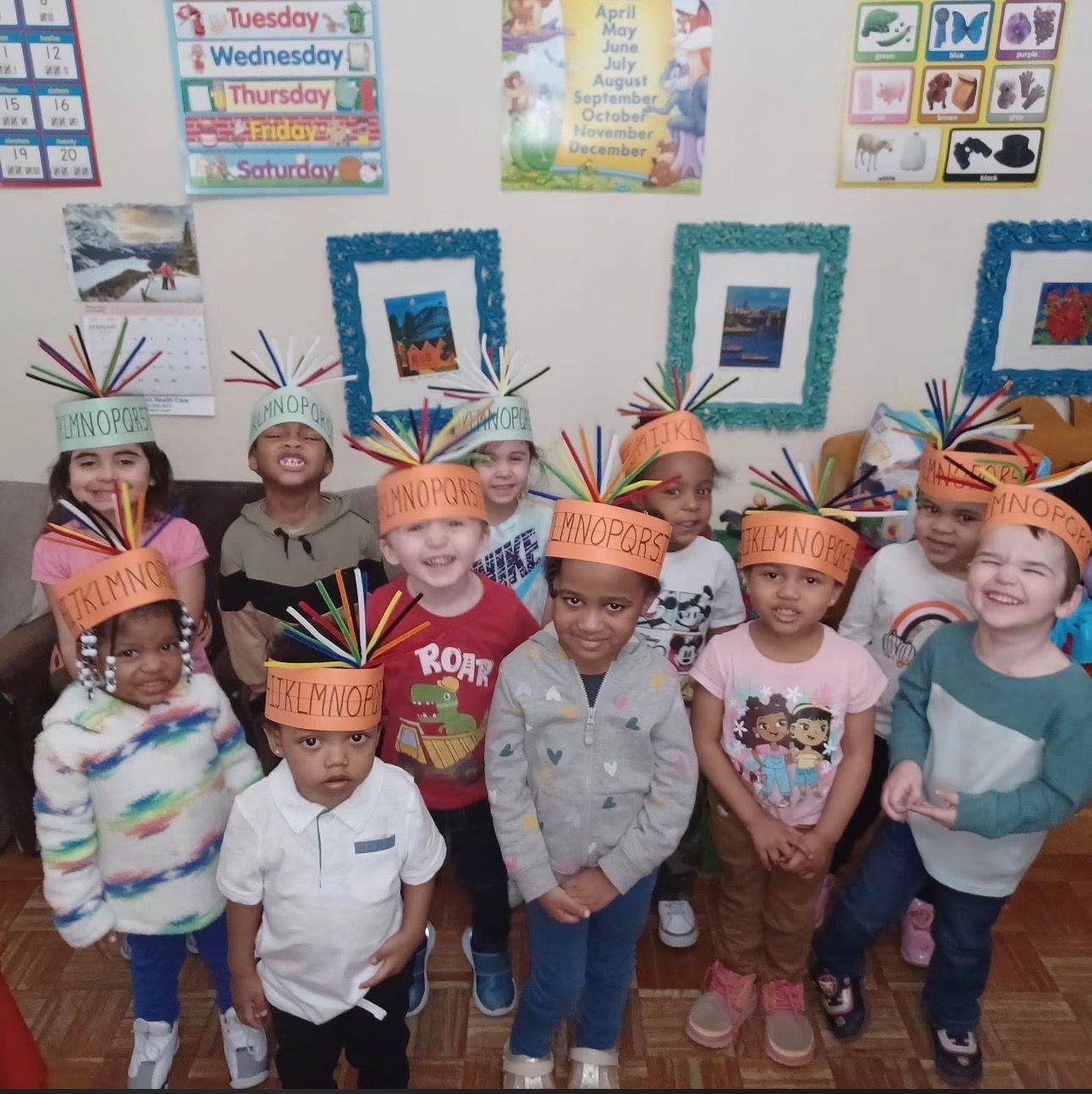 Children in a classroom wearing colorful feather headbands and orange paper hats, smiling for a group photo