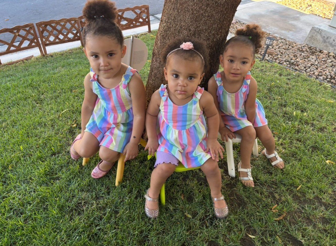 Three toddlers in pastel dresses sitting on grass outdoors near a fence