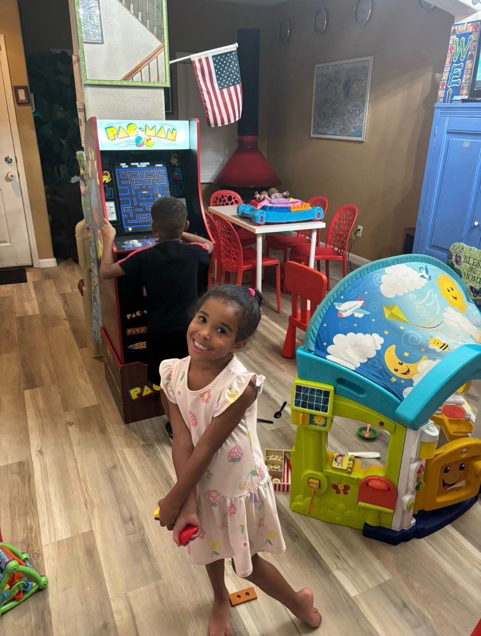 Child posing on a hardwood floor in a playroom, with toys, a small table, and a globe toy nearby.