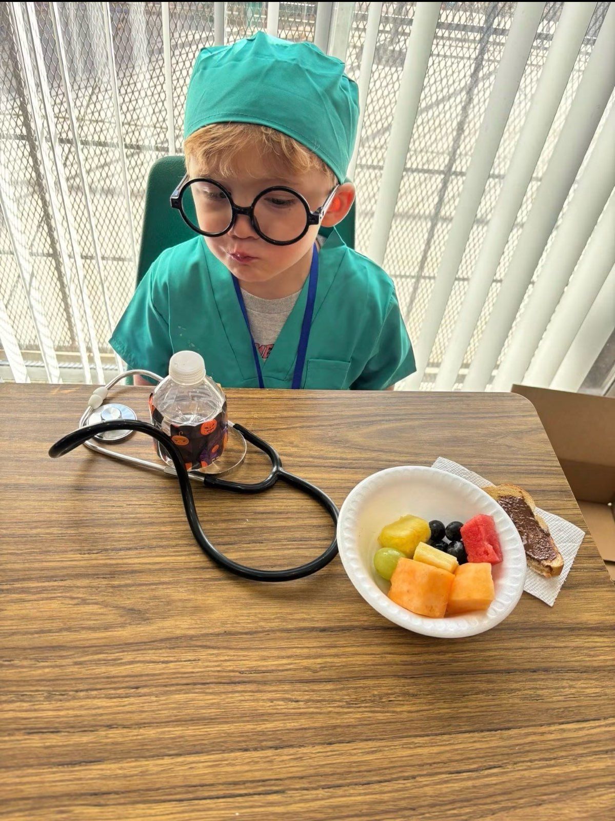 Child in teal surgical scrubs and cap sits at a table with a stethoscope and a bowl of fruit, looking down.