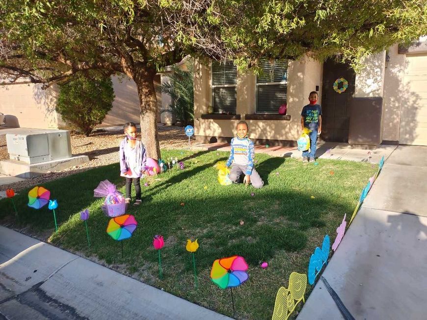 Three children sit and stand in a front yard with colorful pinwheels and a house in the background.