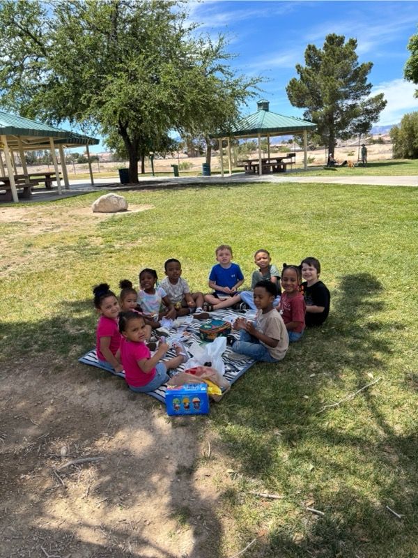 Children sitting on a picnic blanket in a park, gathered around a snack tray under trees.